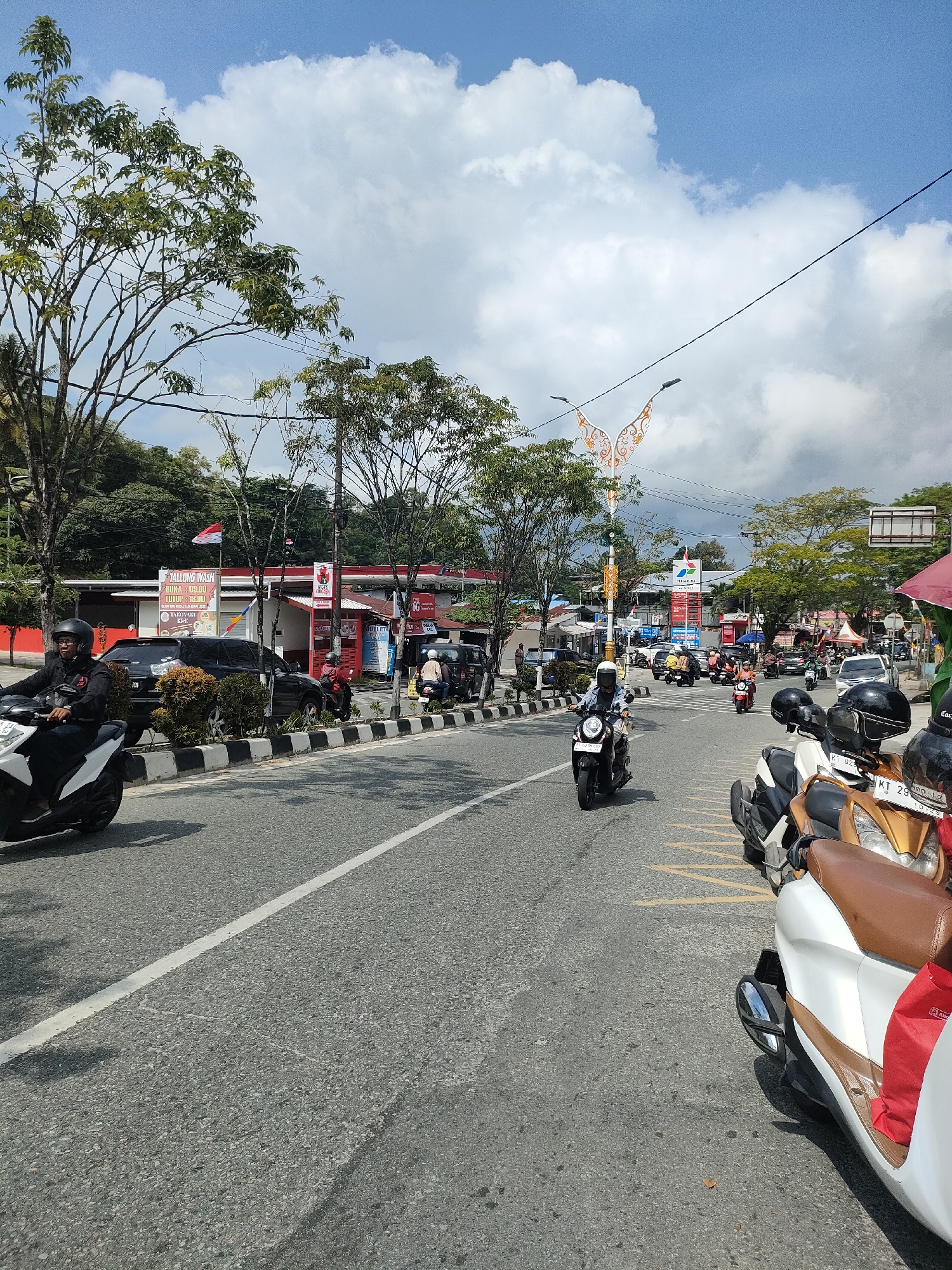 In an outdoor, daytime scene in Unknown, Unknown, a street with multiple lanes of asphalt is lined with trees and buildings. The sky is partially cloudy with blue patches. Several motorcycles are in motion on the road, with riders wearing helmets. On the side of the road, parked motorcycles and cars are visible, alongside buildings that appear to be shops or businesses, including one with a "YALLONG WASH" sign. A red Indonesian flag is displayed on a pole near one of the buildings. The road has white lane markings and some yellow markings near the parked vehicles. The overall impression is of a busy street in a developed area.