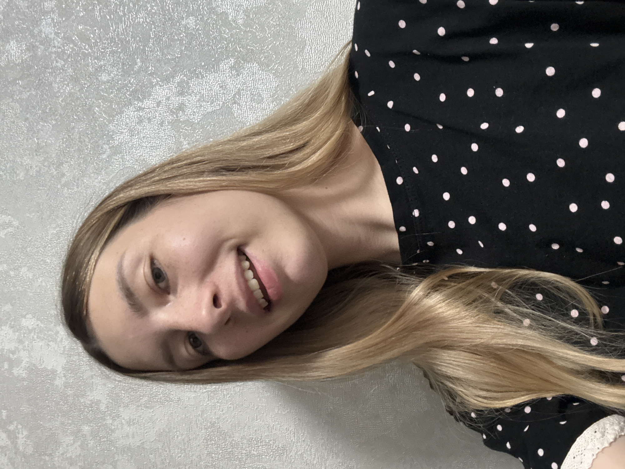 A woman is posing for a picture. She has blonde hair and is wearing a black shirt with pink polka dots. The background appears to be a silver textured wall. There is no discernible evidence to place the location in Moscow, Russia.