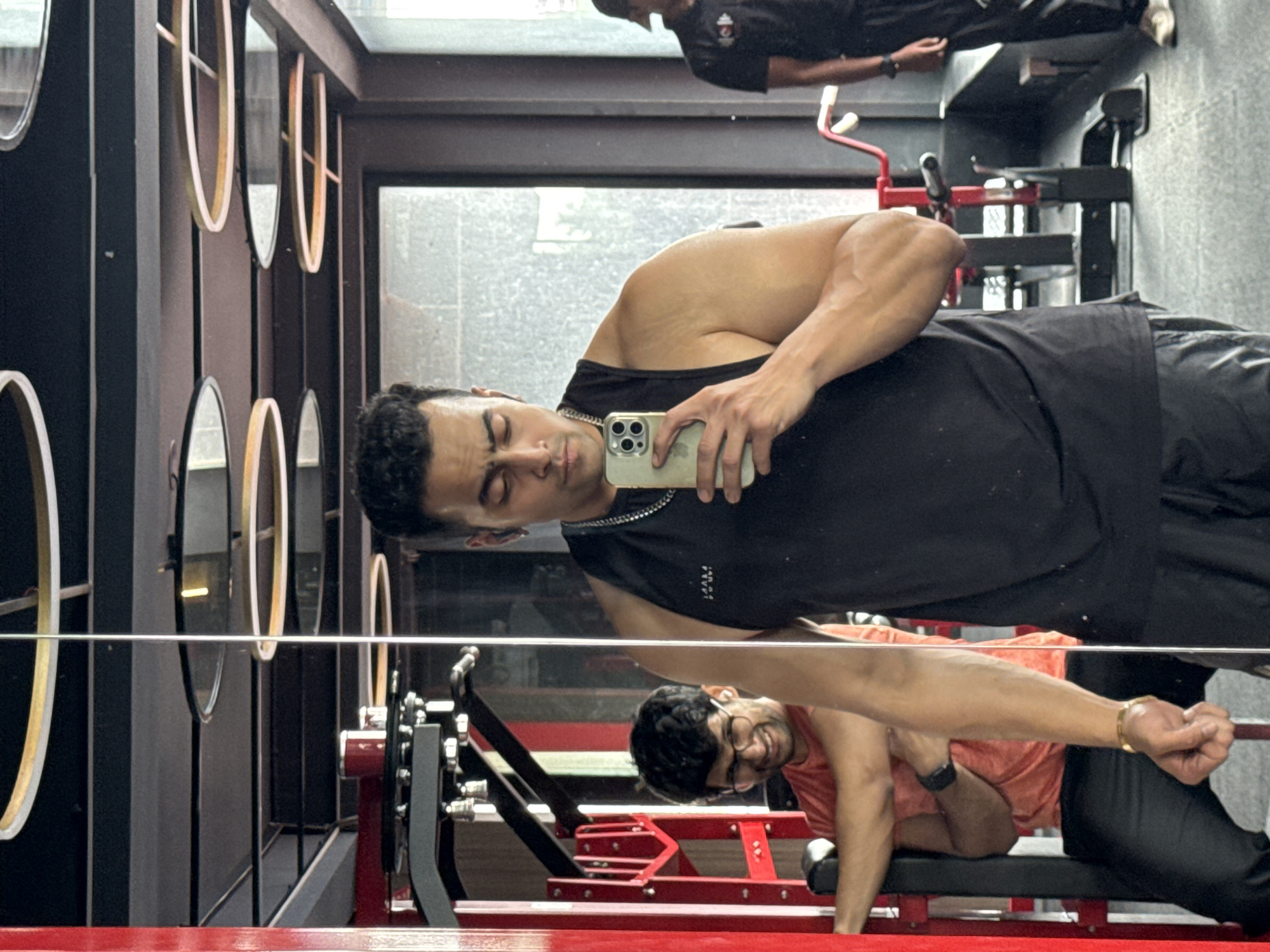 The image shows a man in a black tank top taking a selfie in a gym. He is holding a smartphone in his right hand and looking down at it. He is wearing a silver chain necklace and appears to have a muscular build. In the background, reflected in a mirror, are other gym equipment and people. To the left, another man in an orange t-shirt and glasses is visible, smiling. To the far right, another person in a black shirt and a cap is standing. The gym appears to have modern lighting fixtures on the ceiling. The presence of gym equipment and the overall ambiance suggest an indoor fitness facility. The question specifies Kolkata, India, and given the context of a gym and diverse individuals, it is plausible that this scene is taking place in a gym in Kolkata.