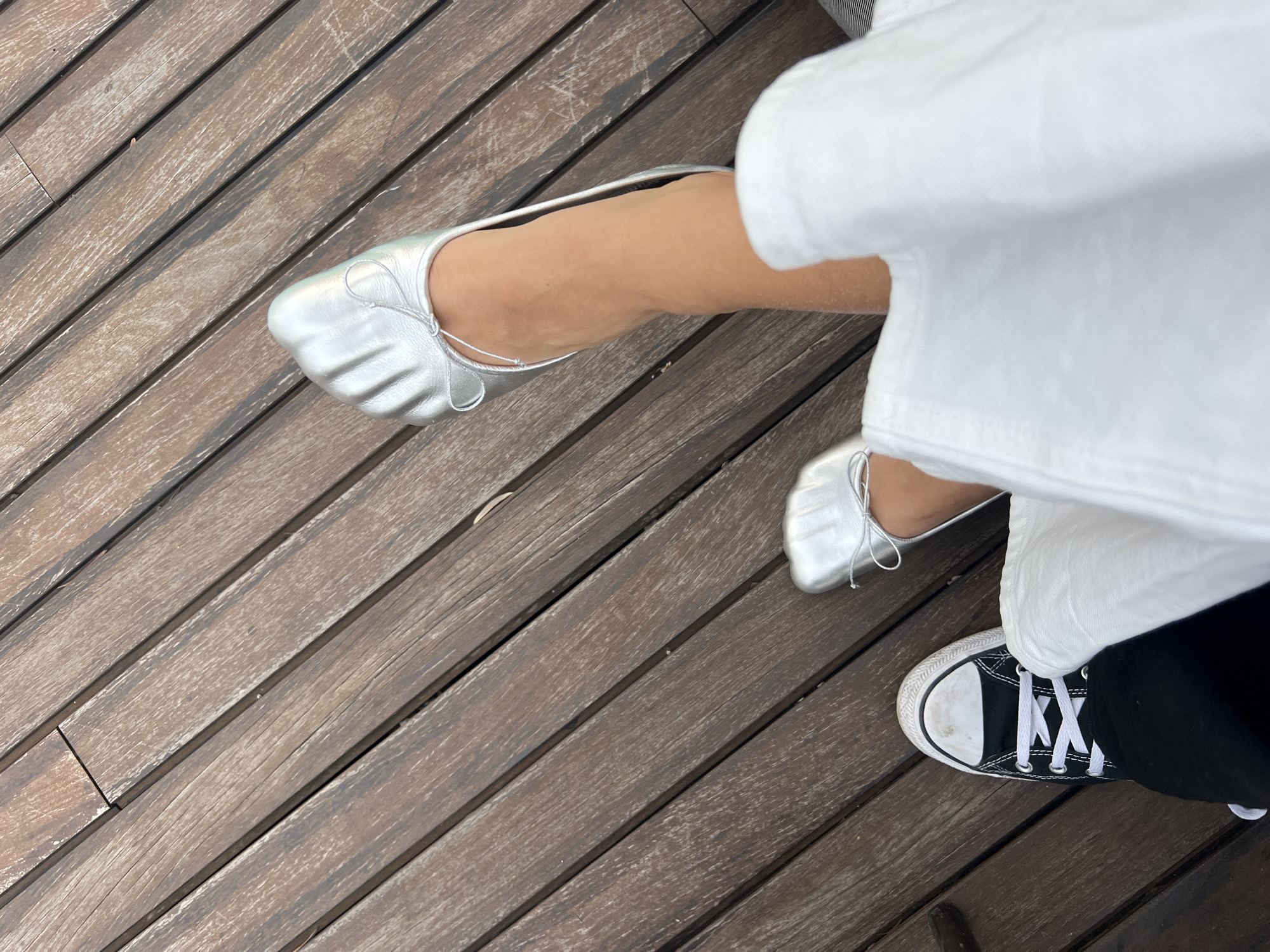 This overhead shot captures the feet of two individuals resting on a weathered wooden deck, likely an outdoor setting in Milan, Italy. The bright, natural light suggests it is daytime.
On the left side, a person's left foot is prominent, adorned in a metallic silver ballet flat with a gathered toe and delicate lacing across the top. The skin of the foot and lower leg appears bare and lightly tanned. Further into the frame, a portion of the other foot, also in a matching silver ballet flat, is visible beneath a flowing white fabric, possibly a dress or wide-leg trousers.
On the right, another individual wears a black Converse-style sneaker with white laces and a white rubber toe cap, showing some wear. This foot is clad in black trousers, with the same white fabric from the first person's attire partially obscuring the top of the sneaker and leg.
The wooden deck features long, narrow planks arranged diagonally across the frame. The planks are dark brown with visible grain and variations in texture, suggesting age and exposure to the elements. No text is visible in the image. The scene conveys a casual, relaxed moment shared between the two individuals.