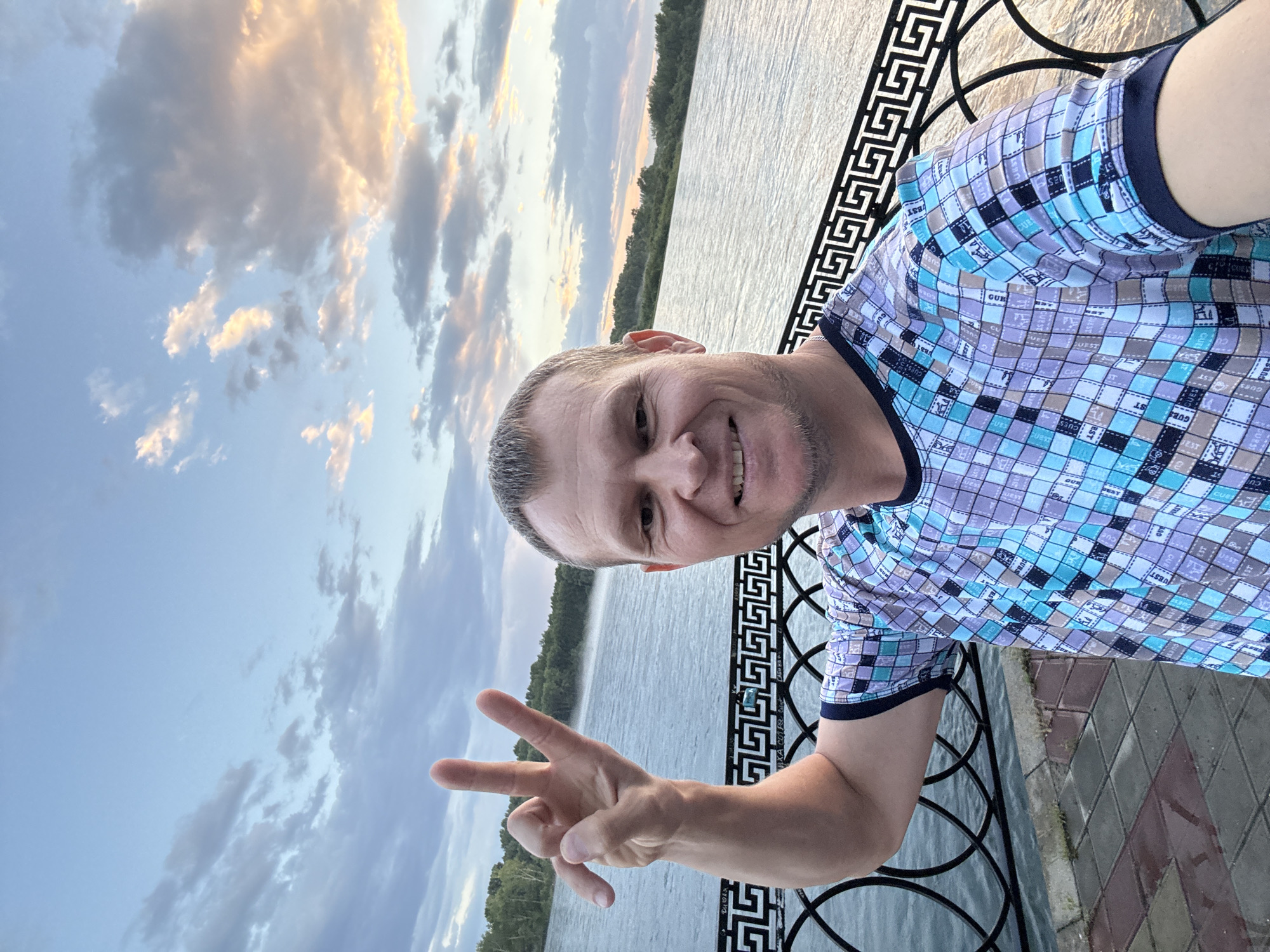 A man with short brown hair and a light beard is taking a selfie outdoors. He is wearing a short-sleeved t-shirt with a checkered pattern in shades of blue, purple, and gray. His right arm is raised, and he is holding up two fingers in a peace sign. He is smiling at the camera.

He is standing on a paved walkway next to a black metal railing with a Greek key pattern. Beyond the railing is a body of water, likely a river, with trees lining the far bank. The sky above is a pale blue with scattered clouds tinged with the colors of sunset. The overall scene suggests a pleasant evening by the water.

This scene appears to be located at the embankment of the Irtysh River in Oskemen, Kazakhstan.