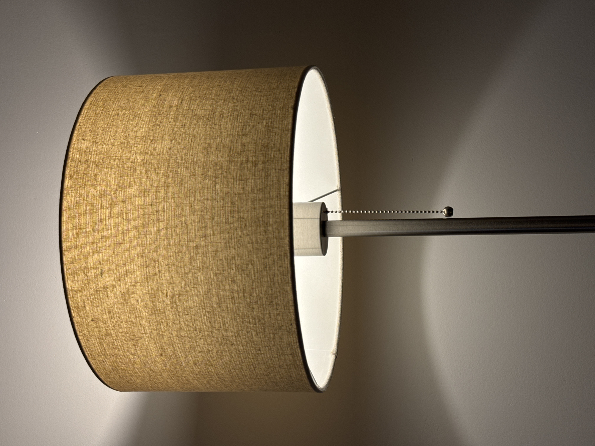 The image shows a close-up of a floor lamp with a textured, woven lampshade in a neutral beige color. The lamp is lit from within, casting a warm glow that illuminates the inside of the shade and projects light upwards onto a plain wall. The wall appears to be painted a light, neutral color. Below the lampshade, the metallic pole of the lamp is visible, along with a pull-chain with a metallic sphere at the end. The scene is dimly lit, with the primary light source being the lamp itself. No people are visible in the image. The photograph is a still shot, capturing a static moment. The location in Bratislava, Slovakia, cannot be determined from the visual information provided in the image.