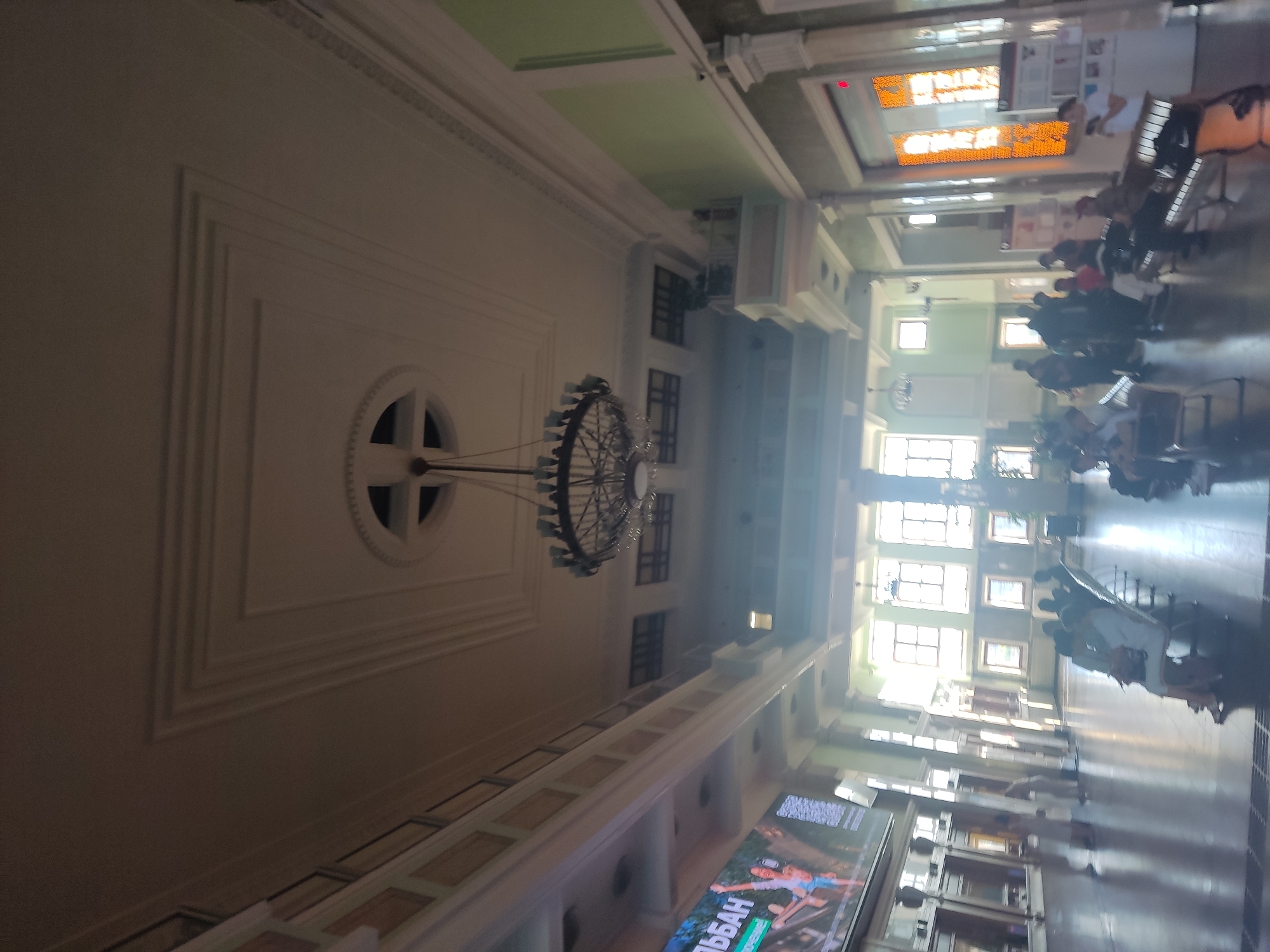 This media file shows a wide-angle view of a grand interior, likely a train station or large public building, in Novosibirsk, Russia. The perspective is tilted, giving an impression of looking up or down along a long hall.
**Subjects:** The primary subjects are the architectural elements of the building, including ornate white moldings on the walls and ceiling, a large, intricate chandelier suspended from above, and rows of windows that admit bright, natural light. Scattered throughout the space are people, mostly seen in silhouette or as blurred figures, sitting on benches or walking. They appear to be waiting or moving through the station. In the foreground, a large digital display screen is visible, showing an advertisement with people in action poses.
**Activity/Event:** The scene depicts ordinary activity within a public transit hub. People are waiting for trains, traveling, or passing through. The atmosphere appears calm and functional.
**Notable Details:** The time of day is difficult to determine precisely due to the interior lighting, but the bright light coming through the windows suggests daytime. The presence of benches, a large hall, and what appears to be a departure board (the orange illuminated display at the top) strongly indicates a train station. The architecture itself suggests a historical or important building, possibly from the Soviet era given the location. The overall mood is one of spaciousness and a steady flow of public life.
**Visible Text:** The most prominent visible text is on the digital display in the foreground. Part of it reads "ЭЙТБАН" (EITBAN), which is likely part of an advertisement. There are also some smaller signs and posters visible in the background, but the text is not legible.