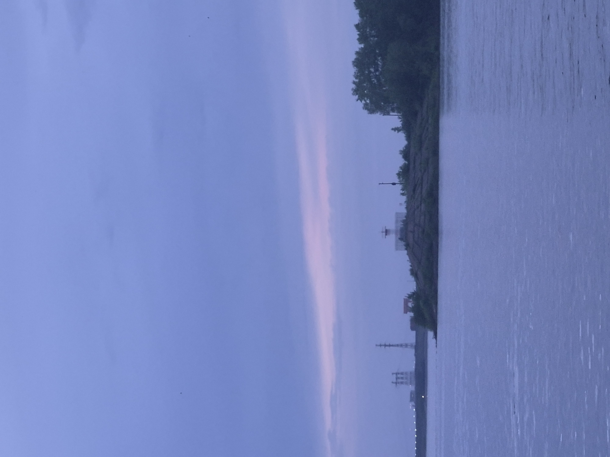 The image shows a body of water on the right, with the opposite bank visible. On the bank, there is a line of trees and what appears to be some type of industrial structure in the distance. The sky is a gradient of blues and pinks, suggesting either dusk or dawn. The location is Unknown, Unknown.