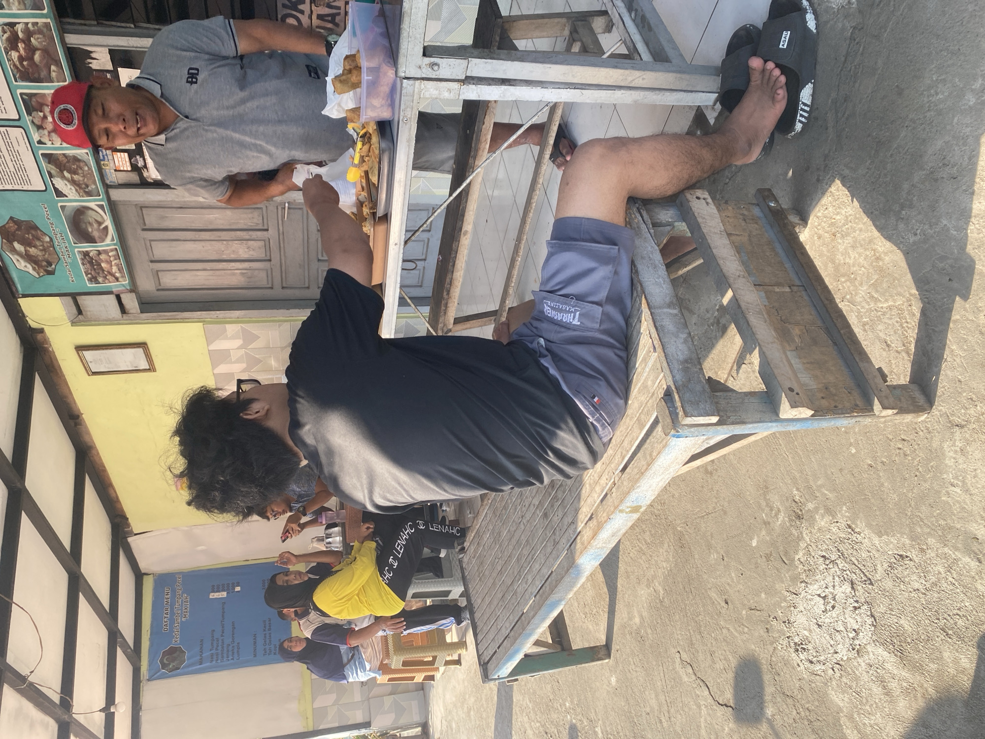 In Kediri City, Indonesia, an outdoor food stall features a man in a red cap and gray polo shirt serving food to customers. He is standing behind a metal table laden with various fried foods, including what appear to be fritters and spring rolls, arranged on metal trays and in a plastic container. In front of the table, a person wearing a black t-shirt and gray shorts is seated on a wooden bench, reaching out to receive food. Further back, several other individuals are seated at a table, with some appearing to be eating and others conversing. A blue banner displaying a menu and "DAFTAR MENU" (Menu List) in Indonesian is visible on the wall behind them. The ground is paved with concrete, and shadows suggest it is daytime.