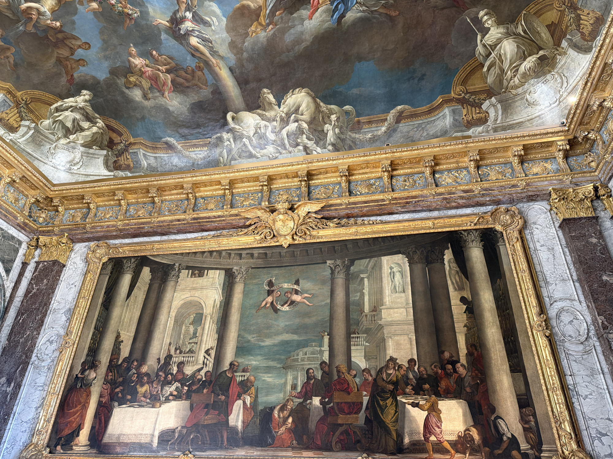 The image depicts the interior of a room in the Palace of Versailles, France, featuring an ornate ceiling fresco and a large framed painting. The ceiling fresco is a trompe-l'oeil painted in blues, golds, and earthy tones, with figures of mythological or allegorical beings in clouds, including horses, chariots, and draped human forms. The fresco is framed by elaborate gilded moldings and decorative elements.
Below the ceiling, a large, rectangular painting is centrally displayed. The painting is an oil on canvas, rendered in rich, deep colors. It portrays a biblical scene, likely "The Wedding at Cana" or a similar banquet. The scene is set in a grand architectural space with towering Corinthian columns and arches, suggesting a palatial or temple-like setting. A long table is laden with food and drink, and numerous figures are seated or standing, engaged in conversation and feasting. In the foreground, figures in period attire are depicted, some in act of serving. Above the central part of the banquet scene, two cherubic figures with wings are depicted in the sky, one holding a scroll with text. The painting is enclosed in a heavy, gilded frame. The walls of the room appear to be adorned with marble and further gilded ornamentation.
