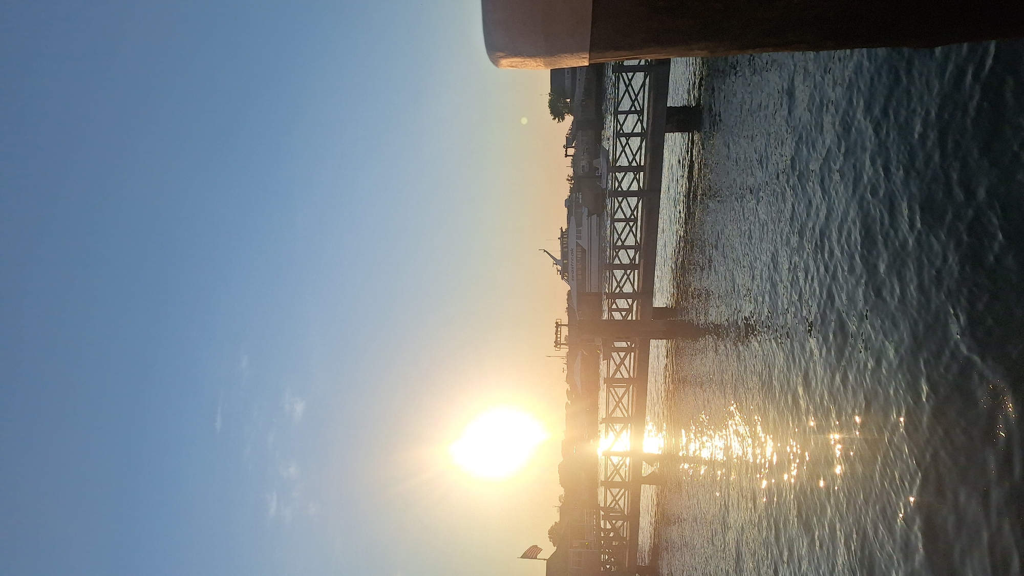 The image depicts a serene waterfront scene at sunset, likely in Nantucket, United States of America, given the architectural style of the buildings and the presence of a ferry labeled "SeaStreak." In the foreground, the calm water reflects the warm, golden light of the setting sun, creating a shimmering effect. A wooden pier with a decorative railing extends across the scene, supported by posts. Beyond the pier, a large white ferryboat is docked, its name "SeaStreak" visible. Several smaller boats are also present in the harbor. In the background, buildings with characteristic New England architecture line the shore, and a flag is visible flying from a building on the left. The sky is a clear, pale blue, transitioning to a warmer hue near the horizon where the sun is setting. The overall atmosphere is peaceful and picturesque.