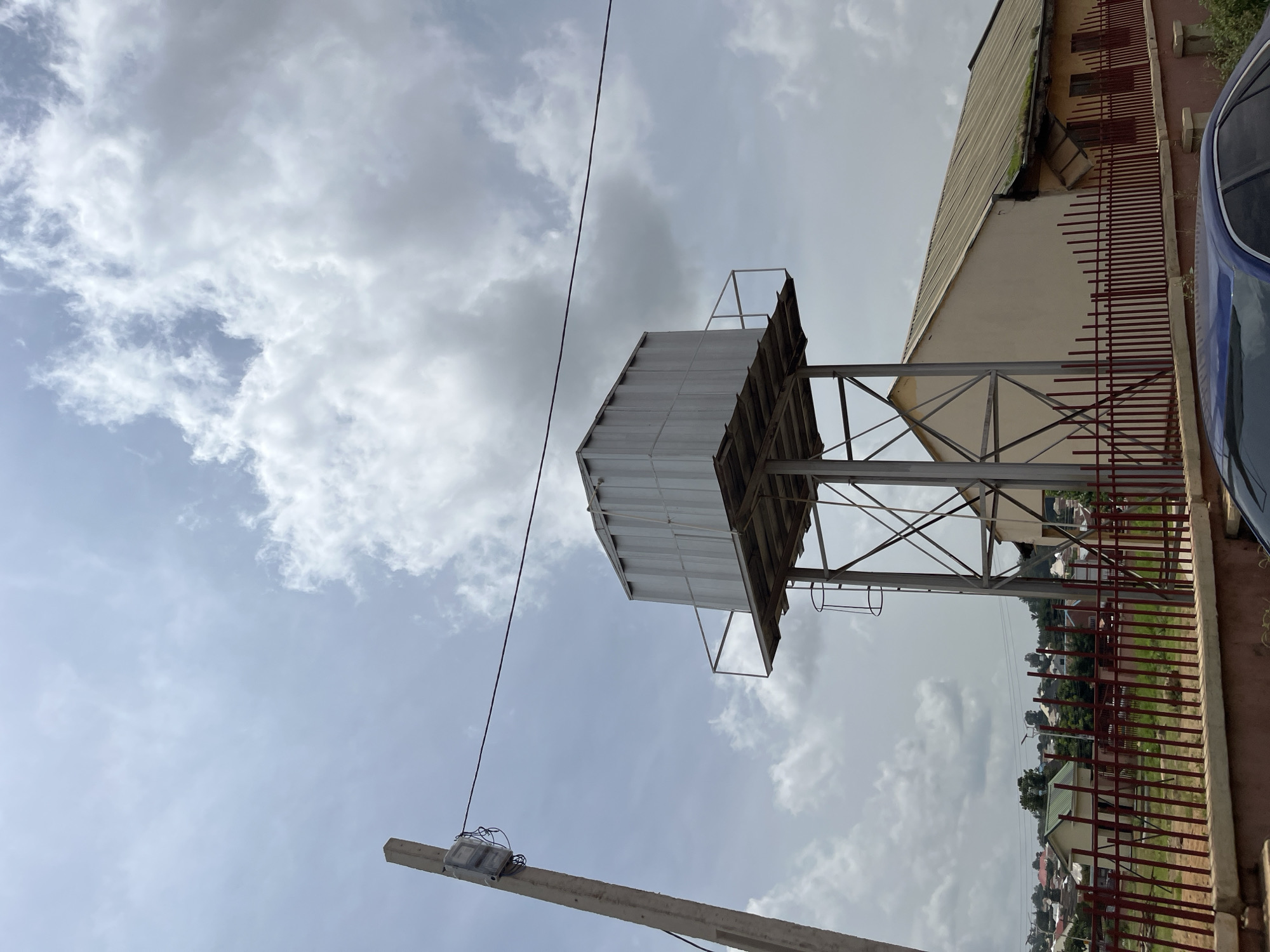 A water tower stands tall in Bukuru, Nigeria, against a cloudy sky. The tower, made of metal, supports a large, silver water tank. A ladder is attached to the side of the tower for access. In the background, a building with a long, sloped roof is visible, as well as houses and trees in the distance. A red metal fence runs along the foreground, and a part of a blue car is visible at the bottom right of the image. A utility pole with electrical equipment is positioned on the left side of the frame.