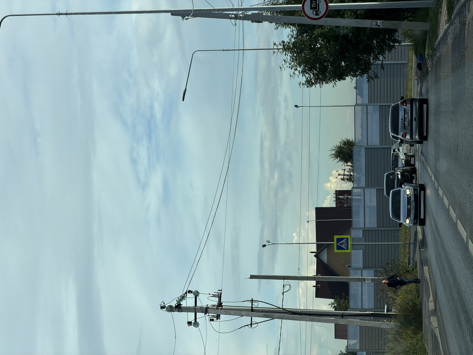The image depicts a street scene with multiple vehicles, a pedestrian, and infrastructure. In the foreground, a gray sedan with license plate K557HC116 RUS is driving on a paved road with dashed white lines. Behind it, another gray sedan is visible, followed by a black SUV. A pedestrian, a man wearing dark clothing, is walking on the sidewalk to the left of the road. He is near a pedestrian crossing sign, which is a yellow diamond with a blue silhouette of people walking.
The road is bordered by a concrete curb and a grassy area with some wild vegetation. Behind the vegetation, a tall, modern sound barrier stretches across the scene, separating the road from what appears to be a residential area. The sound barrier has a textured, metallic surface and is punctuated by vertical supports. In the background, several houses with dark, sloped roofs are visible.
Overhead, a network of power lines and utility poles dominates the sky. The sky itself is a mix of blue and gray clouds. A street lamp is also present on the right side of the image, and a circular traffic sign indicating a weight limit of 15 tons is attached to a pole on the right. The overall environment suggests an urban or suburban setting.
While the image does not contain explicit markers for the city of Kazan, Russia, the Russian license plate on the visible vehicle strongly indicates the location. The architecture of the houses and the design of the sound barrier and utility poles are consistent with the infrastructure found in many Russian cities.