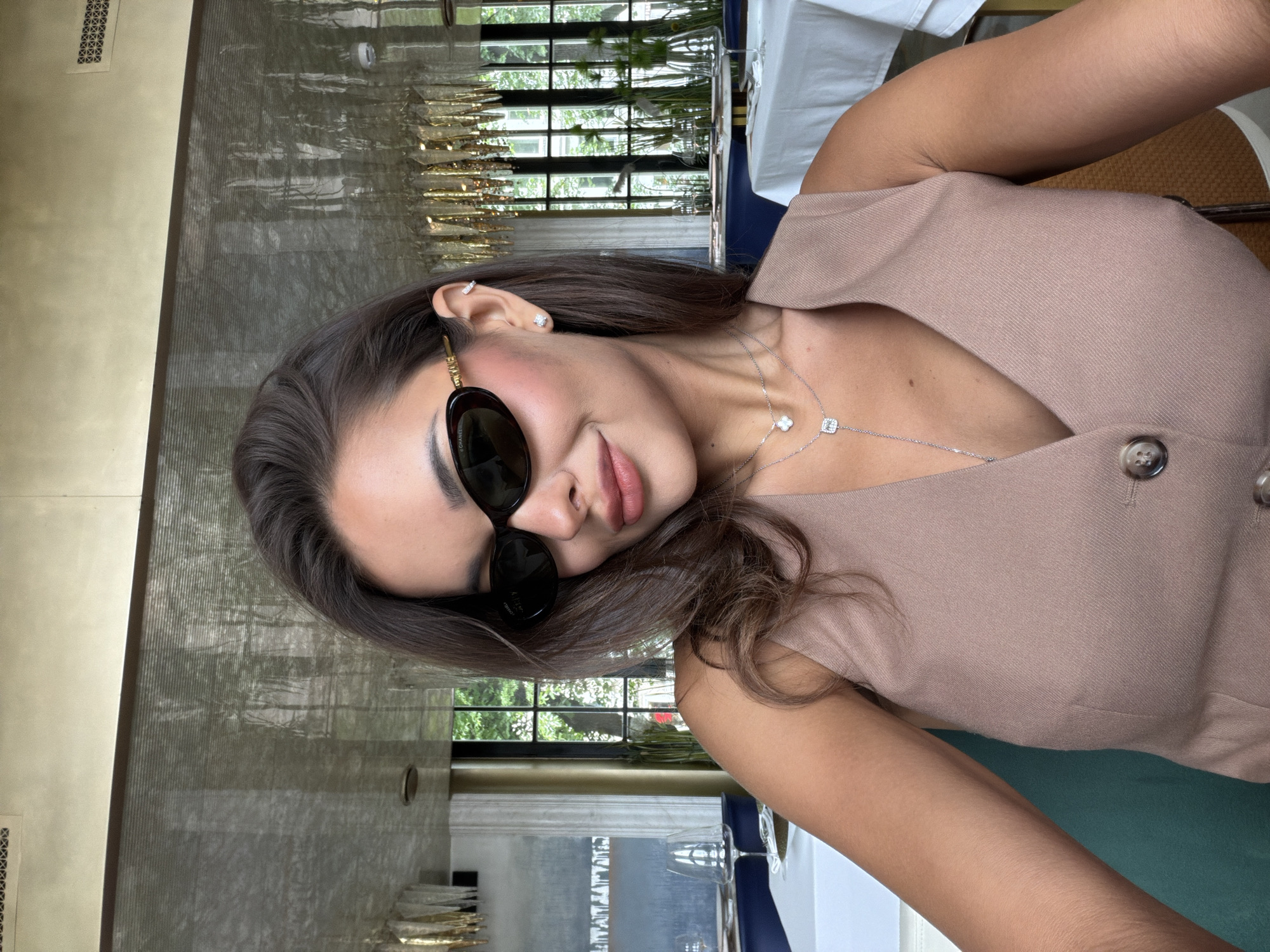 A close-up selfie captures a woman looking confidently at the camera with a slight smile, her head tilted. She has dark hair, styled down, and is wearing large, dark sunglasses with "CHANEL" faintly visible on the arm. Her accessories include a delicate, layered silver necklace with a clover charm and a square pendant, along with sparkling stud earrings and an ear cuff on her right ear. She is dressed in a sleeveless, light brown or taupe top or dress, featuring a textured fabric and visible buttons on the front.
The image is taken indoors, likely within an upscale restaurant or cafe in Moscow, Russia, during the daytime. Behind the woman, large windows reveal a view of green trees and foliage, suggesting a pleasant, warm season. The interior decor features textured, possibly reflective, wall panels. A white tablecloth and a clear wine glass are visible on a table in the background, indicating a dining setting. The overall mood is relaxed and chic.