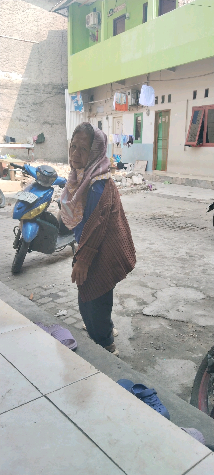 The image captures an outdoor scene in the city of West Jakarta, Indonesia, likely in a residential or informal settlement area. In the foreground, a woman with a hijab, appearing elderly, stands facing forward. She is wearing a brown, ribbed cardigan over a blue shirt and dark trousers. A patterned scarf is draped around her head. To her left, a blue scooter is parked. The ground is paved with concrete and stone, with some scattered debris in the background, suggesting construction or renovation. Behind the woman, a two-story building with a lime green upper facade and white lower facade is visible. Laundry hangs on a line in front of the building. The overall environment appears to be a typical urban setting with signs of everyday life.