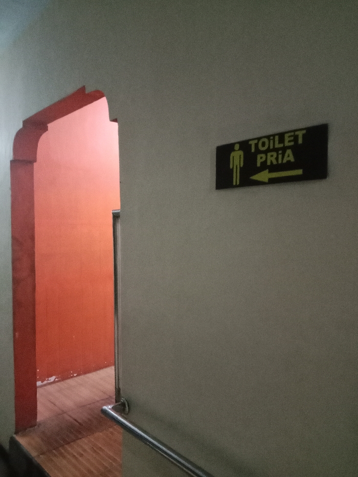 The image shows a hallway with a white wall, an arched doorway with a red frame and an orange wall, and a black sign that reads "Toilet Pria" in yellow lettering. The sign depicts a stick figure of a man and a yellow arrow pointing right. A silver handrail is attached to the white wall. The hallway is located in Tugu, Indonesia, at the coordinates -6.976 latitude and 110.331 longitude. The scene is indoors. No people are visible. The hallway is empty except for the handrail. The image is taken from a low angle. The lighting is dim.