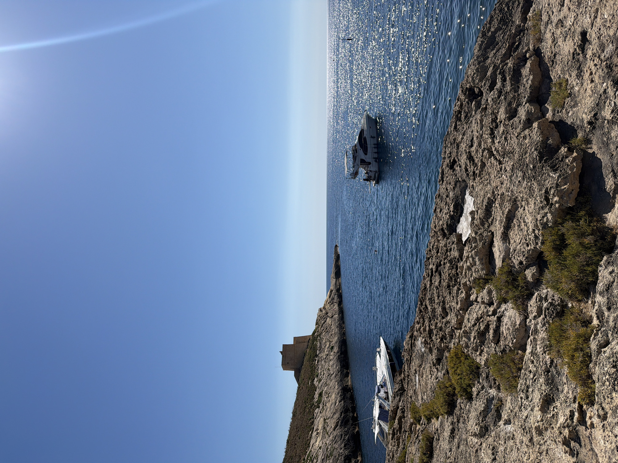 The image depicts a sunny day in Xlendi, Malta, featuring a coastal landscape. The foreground is dominated by rugged, rocky terrain with sparse green vegetation. The midground showcases the deep blue Mediterranean Sea, with two white boats moored near the shore and a larger yacht anchored further out. The yacht has a canopy and several people visible on its deck. In the background, a historical stone tower stands prominently on a hill overlooking the bay. The sky is clear and bright blue, with the sun creating a dazzling reflection on the water's surface.