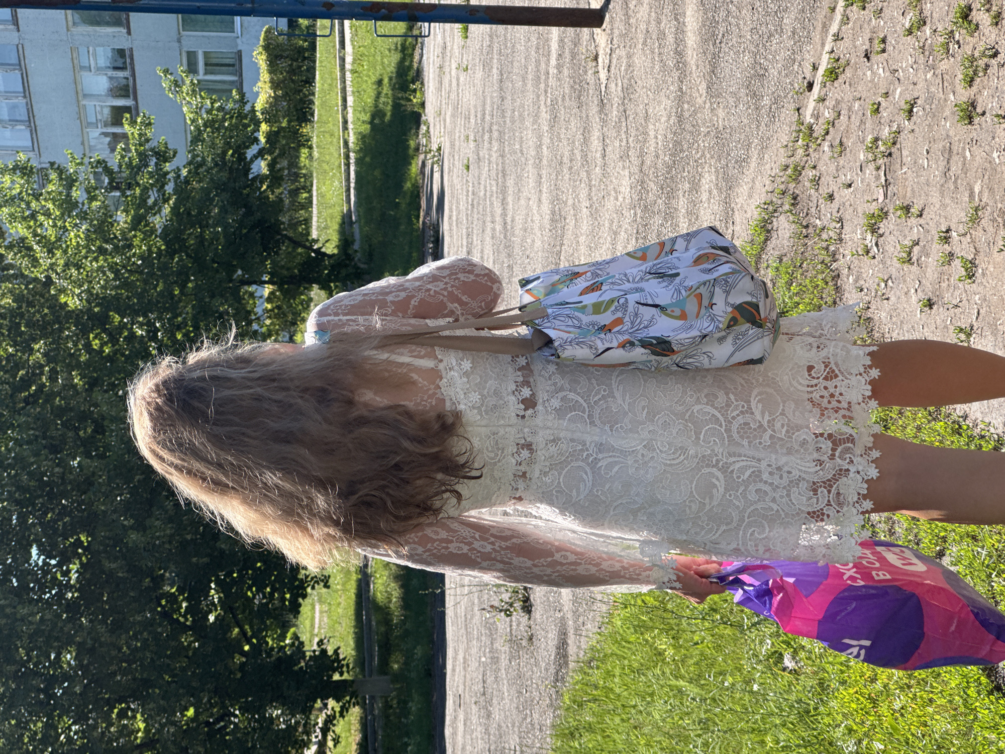 A woman with long, wavy blond hair, wearing a white lace dress, walks away from the camera outdoors. She is carrying a colorful tote bag with a bird pattern on her right shoulder and a purple and pink plastic shopping bag in her left hand. She is on a paved area next to a grassy verge, with trees and a light-colored, multi-story building in the background. The scene is illuminated by sunlight.