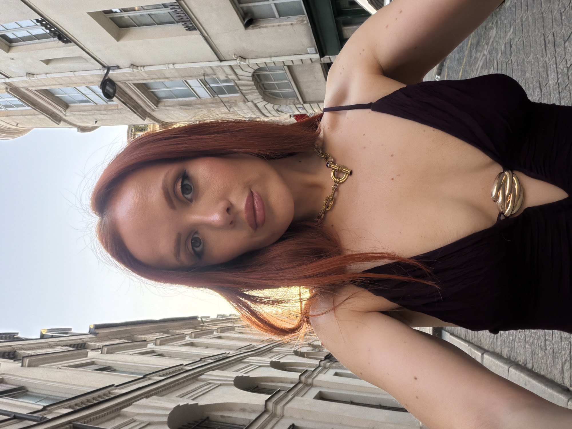 A selfie taken in Paris, France, looking up at multistory buildings. A fair-skinned woman with red hair and a dark-colored dress is centered in the image. She wears a gold necklace. The woman is likely standing on a stone sidewalk. The buildings are light-colored with windows and architectural details. The sky is visible in the background.