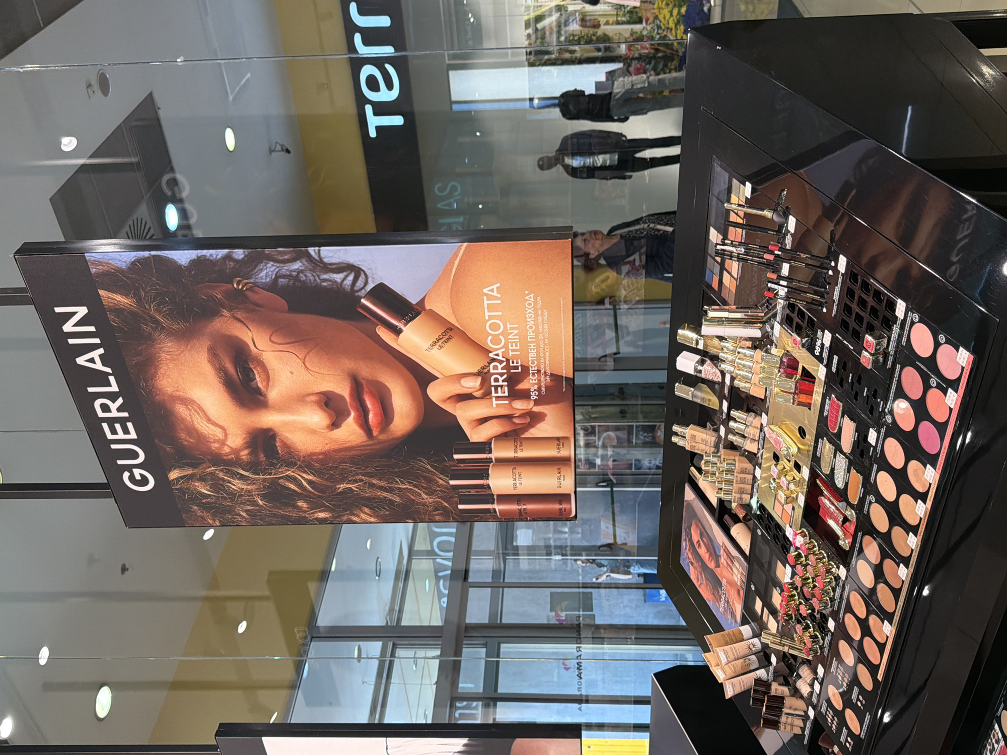 This media file presents a brightly lit cosmetics display within a retail environment in Pleven, Bulgaria. The central focus is a large promotional banner for Guerlain, showcasing their "Terracotta Le Teint" foundation. The banner features a model holding a foundation bottle, with additional product bottles arranged below. Text on the banner, primarily in Cyrillic, states "GUERLAIN TERRACOTTA LE TEINT," and details like "95% ЕСТЕСТВЕН ПРОИЗХОД" (95% Natural Origin), "СЪВЪРШЕНСТВОТО НА ФОН ДЮ ТЕН. ЛЕКОТАТА НА ПУДРА." (The Perfection of Foundation. The Lightness of Powder.), and "24Ч ДЪЛГОТРАЙНОСТ - НЕ ОСТАВЯ СЛЕДИ" (24H Long-Lasting - Leaves No Traces).
Adjacent to the banner is a multi-tiered black display counter filled with a wide range of Guerlain makeup products. These include various shades of liquid foundations in bottles and tubes, eyeshadow palettes, blush compacts, lipsticks, and makeup pencils, all neatly arranged for customer browsing or purchase. Some products also show "98%" on their labels.
The background consists of reflective glass panels, revealing the interior of what appears to be a modern shopping mall or large department store. Reflections show indistinct figures of people, likely shoppers or store personnel, moving about. Partial signage from other stores, such as "Terr" (possibly "Terranova") and "ЯСАМАЯ," is also visible in the reflections. The overall scene depicts a well-stocked and active beauty counter during what appears to be daytime.