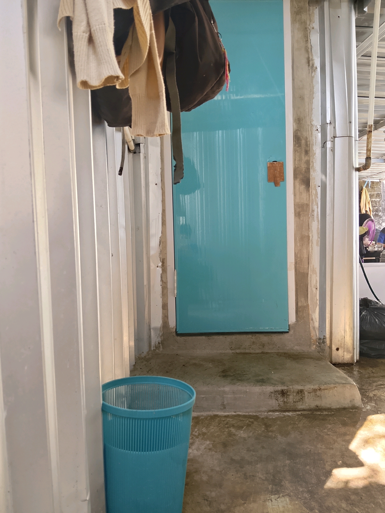 A light-colored garment, possibly a sweater or cardigan, hangs on a hook near a teal-colored door. The garment appears to be made of knitted material and has a textured weave. Next to it, a dark brown backpack is also hanging. The background shows a rough concrete wall and the teal door, which has a small wooden fixture attached to it. To the right of the door, there are vertical corrugated metal panels. In the foreground, a bright teal plastic trash can with vertical ridges stands on a concrete floor that appears wet in some areas, suggesting it might be an outdoor or semi-outdoor space. The sunlight casts a shadow on the floor. The scene is located in Negeri Agung, Indonesia.