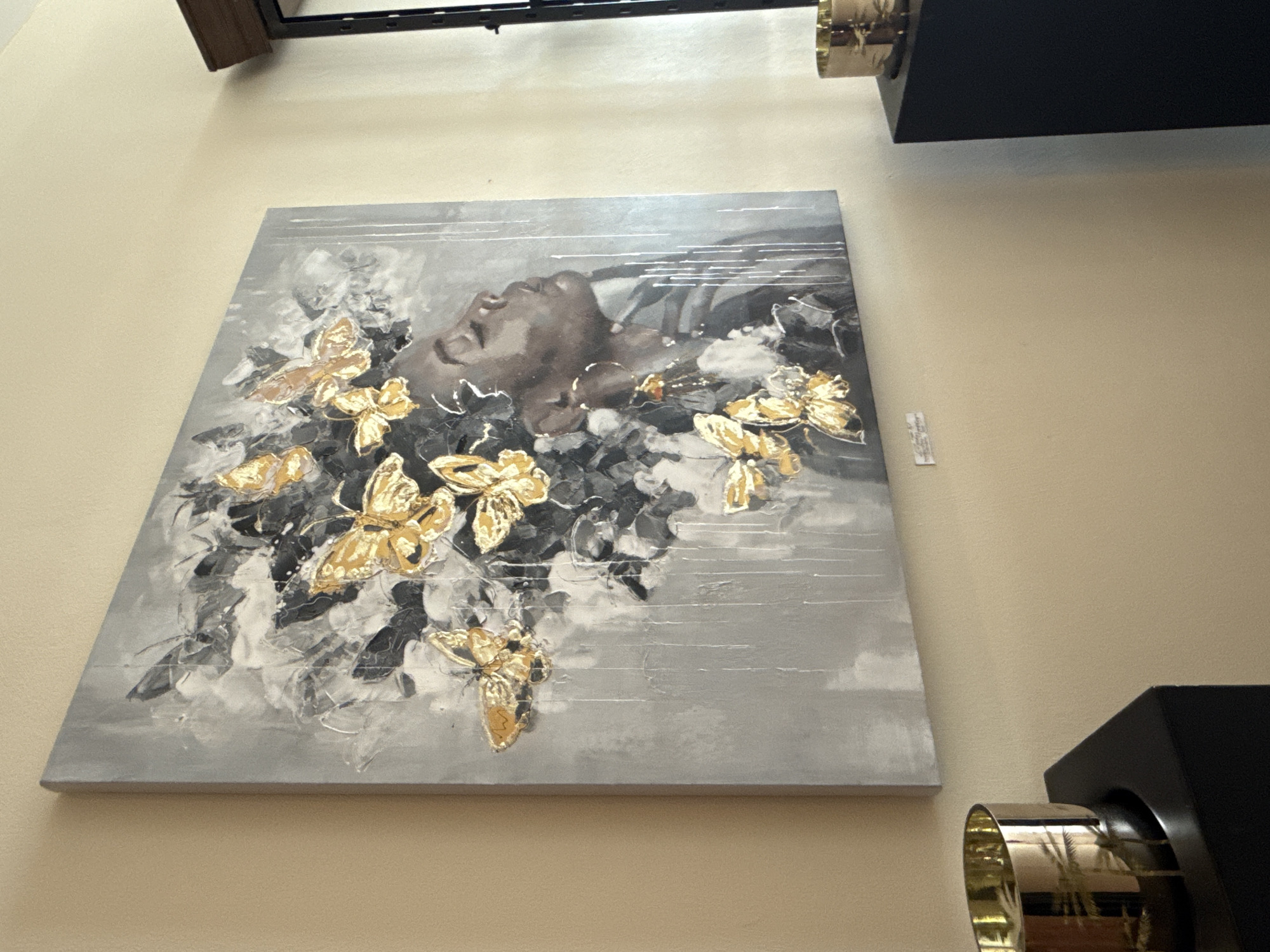 The image shows a large, square painting of a woman's profile with golden butterflies in her hair, hanging on a light beige wall. The painting is abstract and features a dark-skinned woman with her eyes closed, facing to the right. Her hair is depicted as dark, textured, and voluminous, interwoven with numerous golden butterflies. Some of the butterflies are detailed, while others are more abstract. The background of the painting is a textured gray with white and black elements, giving a sense of depth and movement. White, vertical lines are painted across the lower right portion of the canvas.
To the left of the painting, partially visible, is the top of a black pedestal with a brass bowl sitting on it. The bowl is cylindrical with a flared rim and appears to have an etched design of palm trees.
To the right of the painting, also partially visible, is another black pedestal with a similar brass bowl on top. Further to the right, a dark frame with a thin wooden top and a black metal structure is visible, suggesting a window or doorway.
The painting is located in the interior of a room with light beige walls. Based on the context of the provided information, this scene is situated in Bad Sachsa, Germany, specifically within the Kunst- und Galeriehaus (Art and Gallery House) which showcases art by various artists.