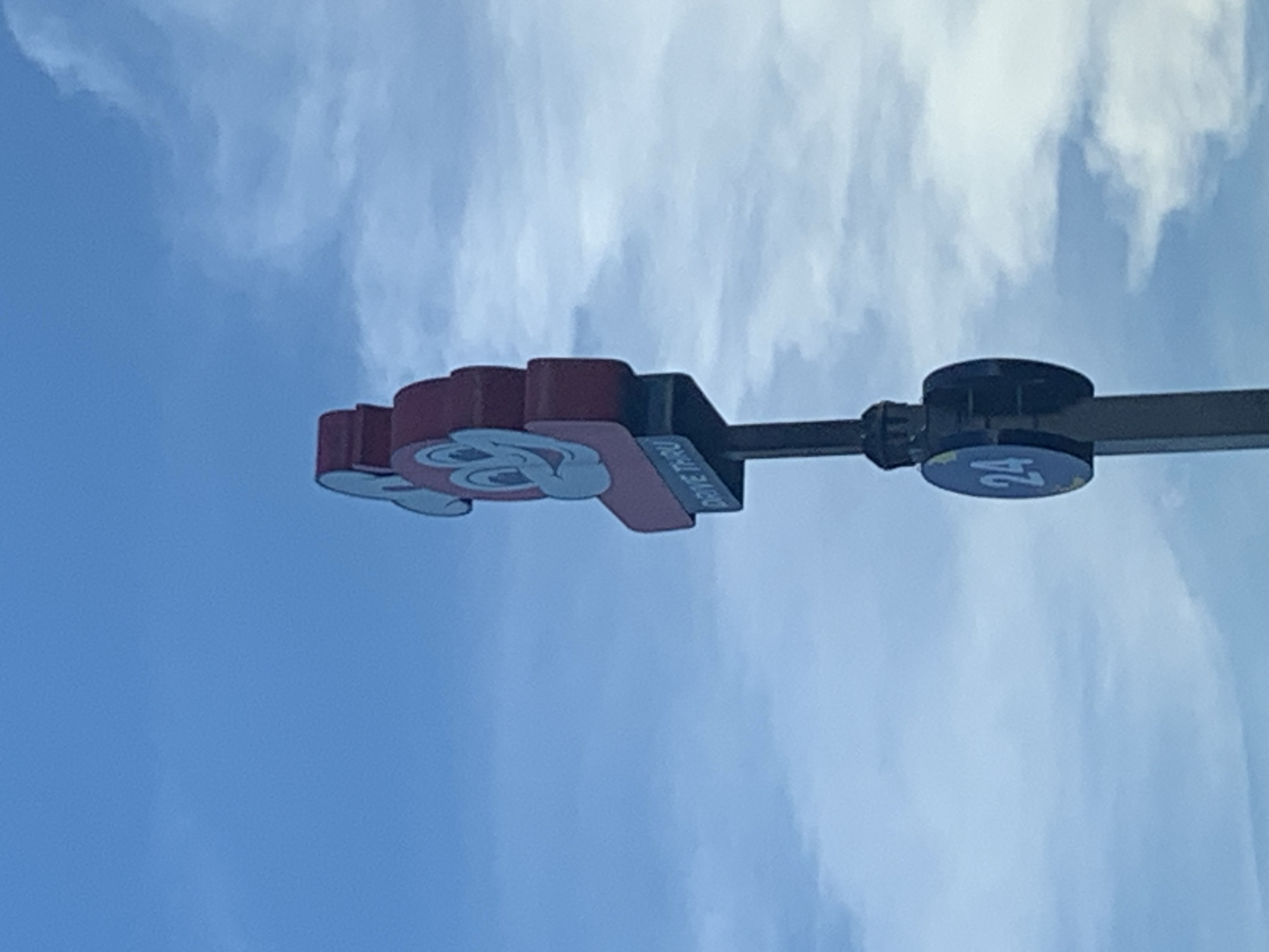 An outdoor, medium shot captures a sign for Jollibee's drive-thru in Angeles City, Philippines, against a backdrop of a hazy blue sky with scattered clouds. The Jollibee mascot, a stylized red bee with a wide smile and light blue accents, is prominently displayed at the top of the sign. Below the mascot, a sign with light blue text on a darker blue background reads "DRIVE THRU." Further down the pole, a circular sign with a dark blue background and yellow accents displays the number "24" in light blue. The pole itself is dark and appears to be metal. The image is taken from a low angle, looking up at the sign.