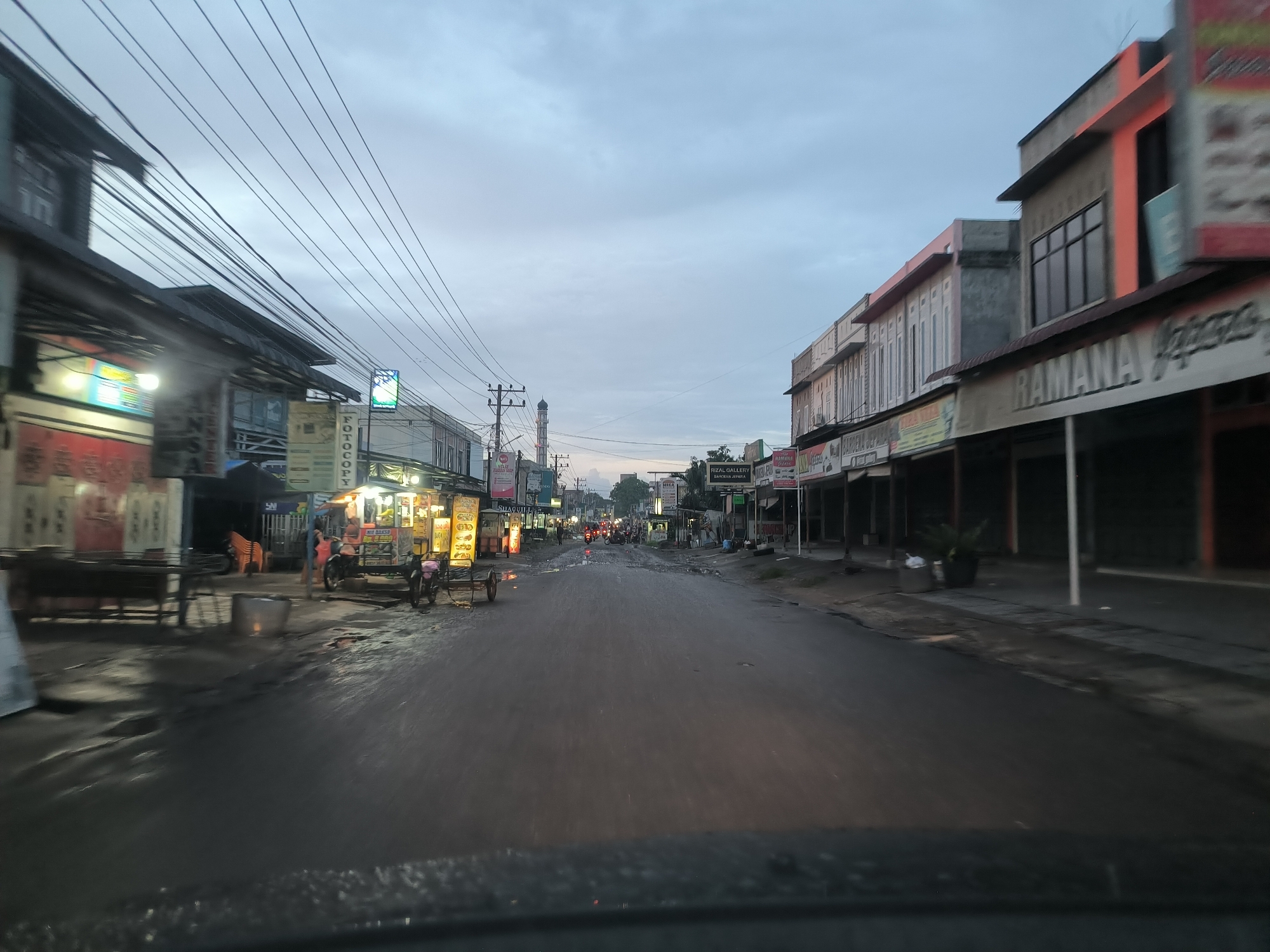 The image captures a street scene in Tambonbaroh, Indonesia, at dusk. The road, appearing wet, stretches into the distance, flanked by two-story buildings that house commercial establishments. Some are closed, indicated by their shuttered fronts. On the left side of the street, food vendors are setting up, evidenced by their carts and lighting. Several people are milling around the vendor area. Power lines crisscross the sky above the street. A mosque minaret is visible in the distance, aligned with the center of the road. The photograph appears to be taken from inside a vehicle, looking out through the windshield.