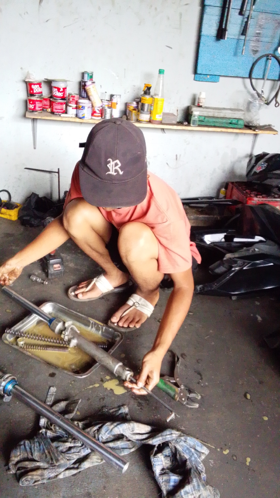 In a dimly lit garage, a person wearing a dark baseball cap and an orange t-shirt is crouched on the concrete floor, surrounded by tools and motorcycle parts. The person is working on a disassembled motorcycle suspension system, with various components laid out in a metal tray filled with oil. A set of pliers and a long metal rod are also present. In the background, shelves are stocked with cans and bottles, possibly lubricants or cleaning supplies. The overall scene suggests a mechanic's workspace, likely in the city of Cintaratu, Indonesia, where repairs are being performed.