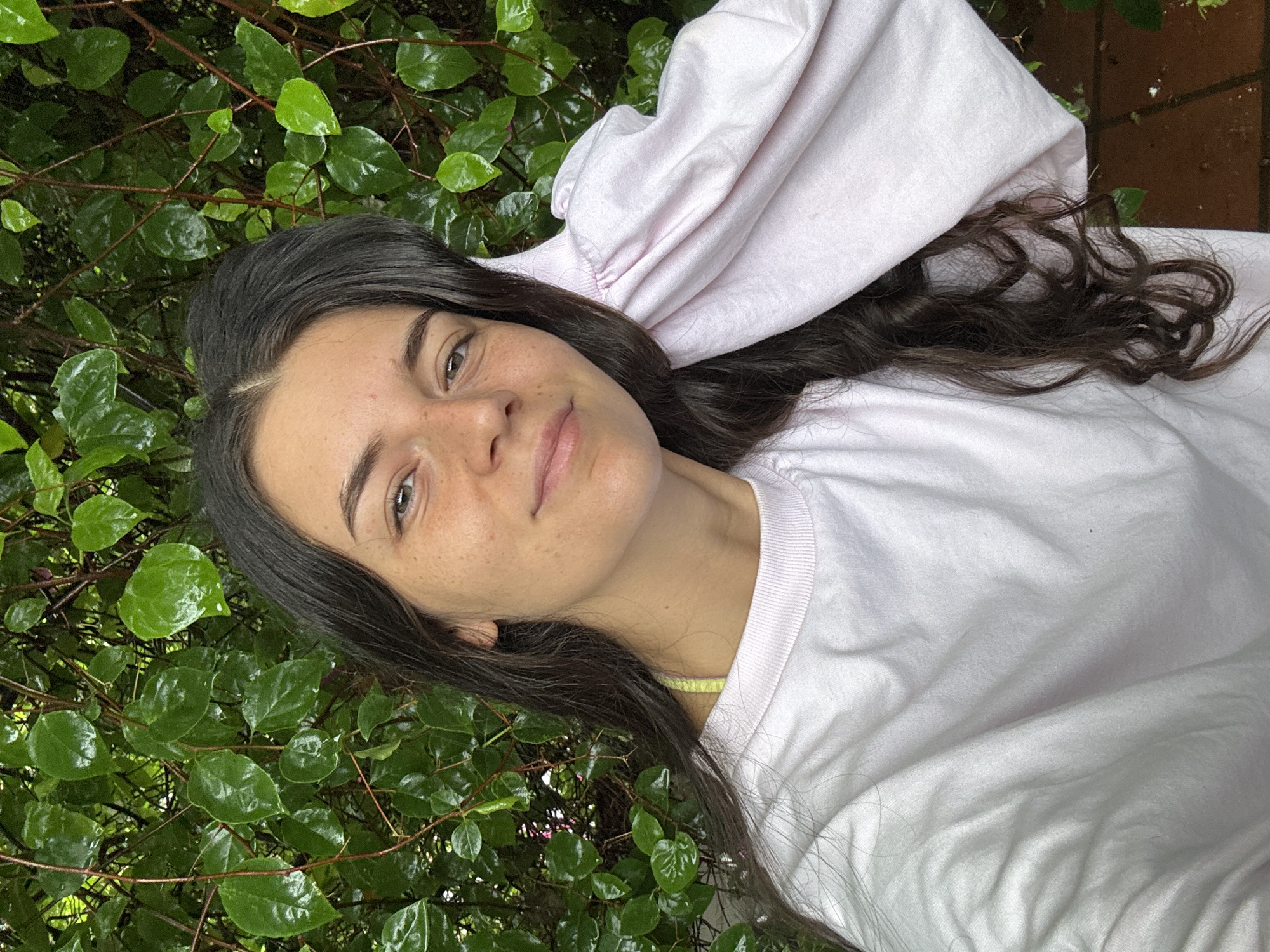 A woman with dark hair and freckles on her face is lying on a pile of lush green leaves. She is wearing a light pink sweatshirt. The location appears to be outdoors, possibly in a garden or park. It is not possible to ascertain the location is in Mũi Né, Vietnam from the image.