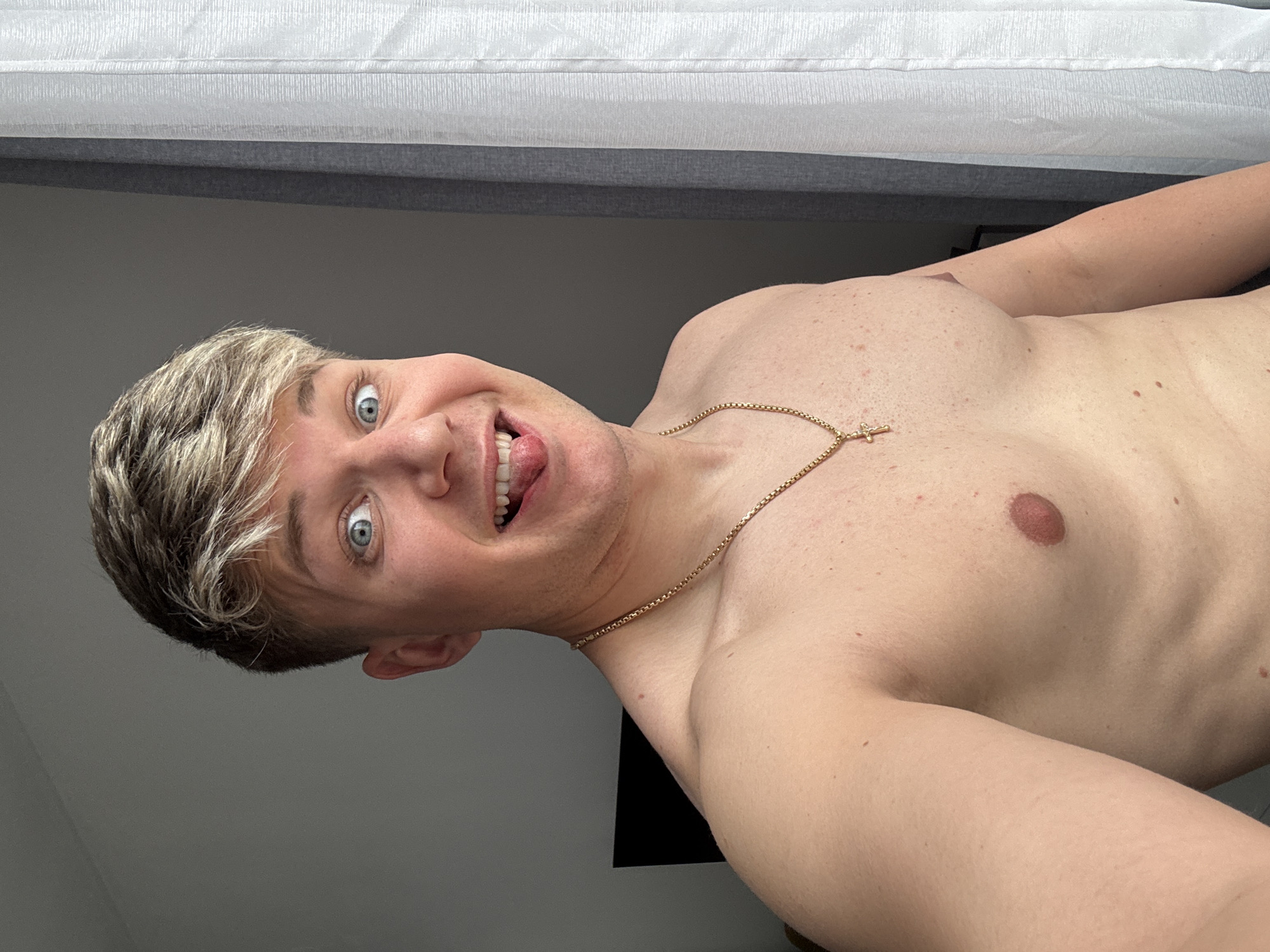 A light-skinned male with blonde hair lies on his back, wearing a gold chain with a cross pendant. His eyes are wide and his tongue is partially out. The background includes a white and gray bed frame and a gray wall. The location is not evident from this image, and there is no information suggesting the image was taken in Plovdiv, Bulgaria.