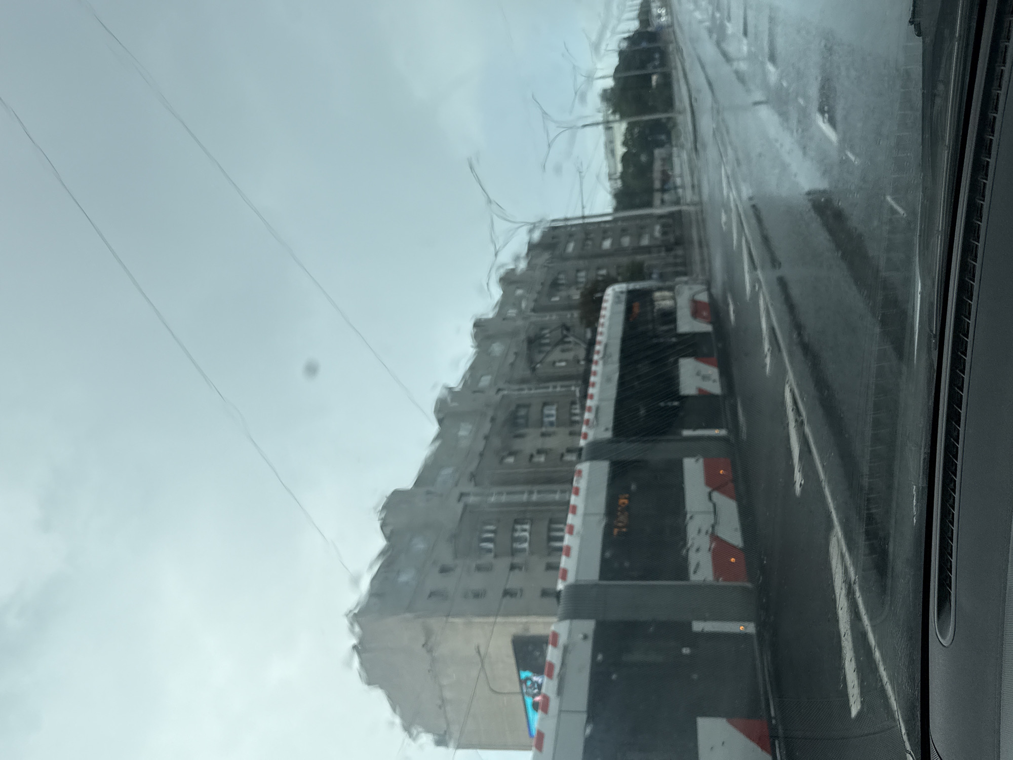 The image shows a street scene in Tallinn, Estonia, on a cloudy and wet day. A tram, partially visible, is traveling along the wet street, which has several puddles. Across the street, a multi-story building with many windows is visible. The sky is overcast, and there are power lines running overhead. The view appears to be from inside a vehicle, looking out through the windshield. The overall impression is of a typical urban scene during inclement weather.