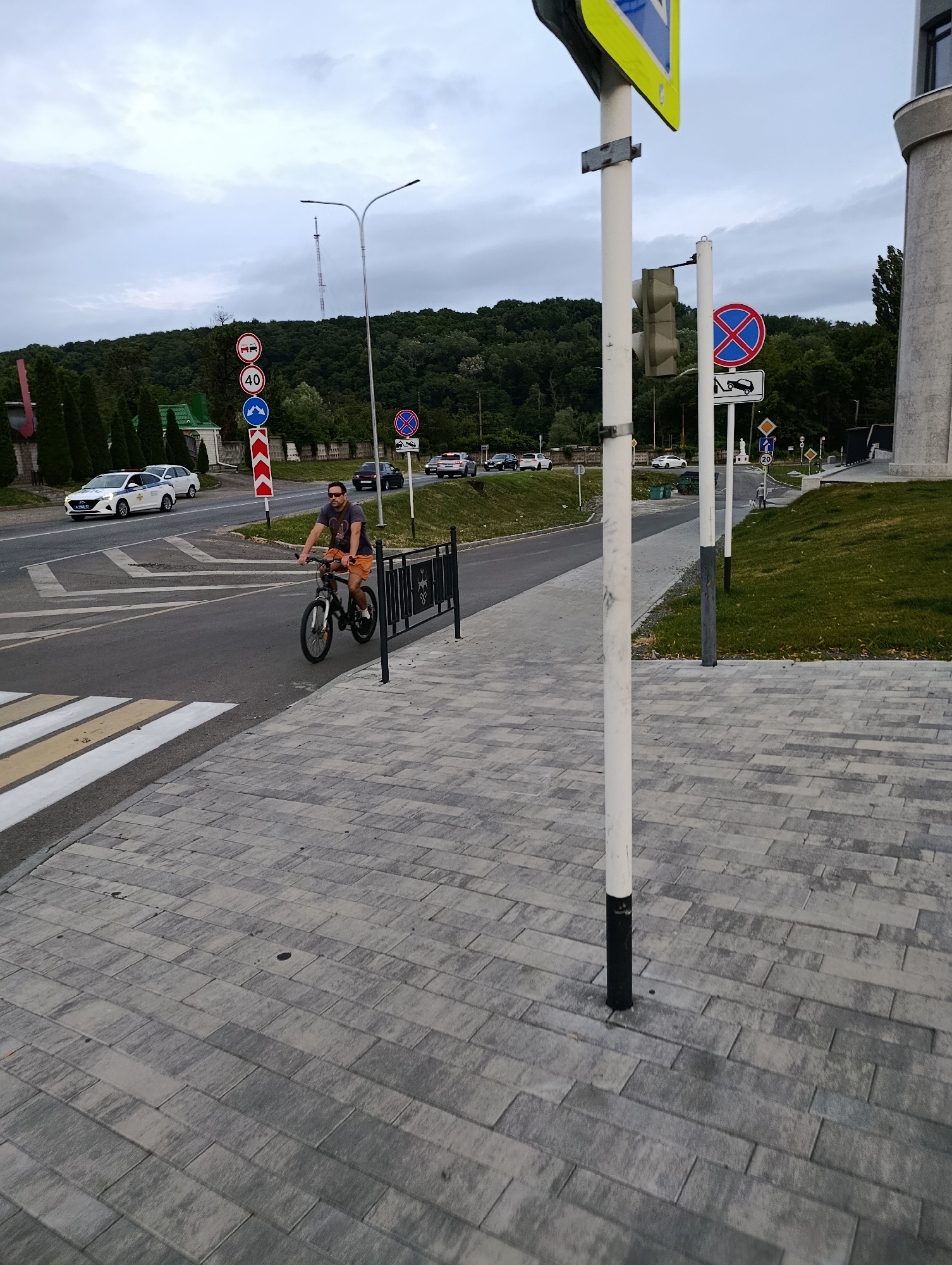 A man is riding a bicycle on a paved sidewalk next to a road. He is wearing a dark t-shirt, orange shorts, and sunglasses. The road has a pedestrian crossing with white and yellow stripes. Several cars are on the road, including a white police car. There are traffic signs, including a speed limit sign (40), a no stopping sign with a tow truck icon, and a left turn only sign. In the background, there are trees and a hill with a telecommunication tower. The sky is overcast. The location is identified as the city of Maykop, Russia.