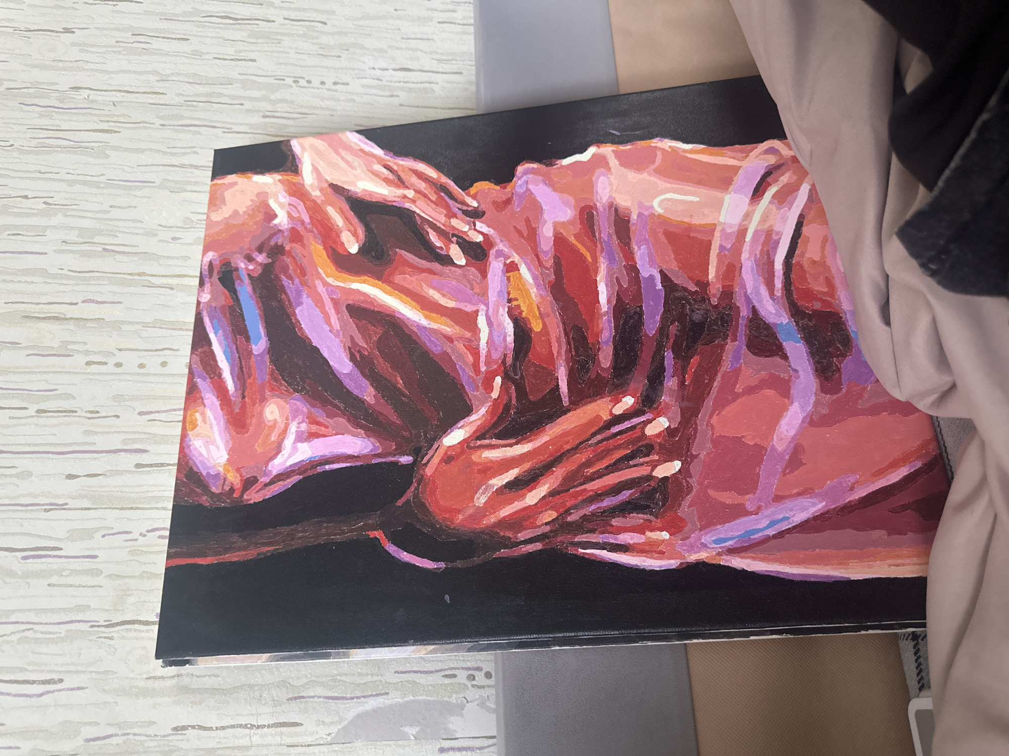 The image shows a painting resting on a surface with a light-colored, wood-grain pattern. The painting features two figures in a reddish-purple color palette against a black background. The figures are intertwined, with hands appearing to reach out. Additional objects, including gray and brown textiles, are visible in the surrounding environment, along with sections of a beige fabric and part of a dark piece of clothing. The photograph's origin location is Высокий, Russia.