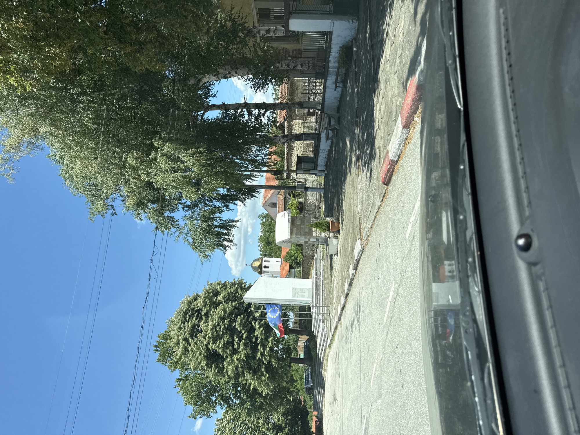 This is a photograph of a village square in Gornoslav, Bulgaria, taken from the perspective of someone inside a vehicle. The focal point of the image is a monument with flags, including the European Union flag and the Bulgarian flag, prominently displayed. Behind the monument, a white church with a golden dome and a cross is visible, surrounded by trees and traditional village houses with red-tiled roofs. The square is paved with interlocking bricks, and a sidewalk with a red and white curb runs along the road. Benches are placed along the sidewalk, and trees provide shade. The sky is clear blue with some wispy clouds. The overall impression is a serene and picturesque village scene on a sunny day.