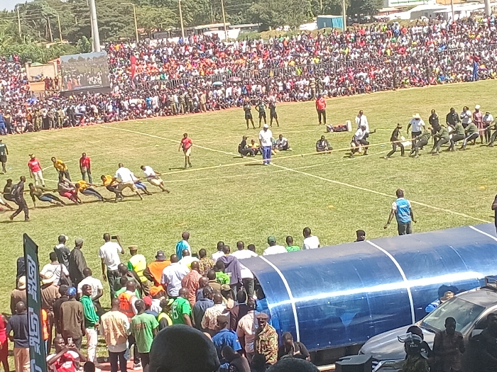 This image captures a vibrant outdoor sports event taking place on a sunny day in Siaya, Kenya. The central focus is a tug-of-war competition occurring on a large, green grassy field. Several teams are actively engaged in pulling on ropes; at least two separate matches appear to be in progress simultaneously. Competitors are dressed in various sportswear, including some teams in coordinated outfits like green or yellow shirts.
A massive crowd of spectators fills the background, occupying bleachers and standing areas, suggesting a highly attended public gathering or festival. The stands are densely packed with people wearing diverse casual clothing. On the far left, a large screen or jumbotron is visible, displaying content related to the event to the distant audience.
In the foreground, a group of people stands behind a transparent blue canopy or shelter, possibly indicating a VIP or designated viewing area. Cars are partially visible to the right of this structure. Scattered yellow cones mark areas on the field, and several officials or referees, some in white shirts, are overseeing the matches. The bright sunlight indicates clear weather and casts distinct shadows across the field. On the left side of the foreground, a tall green banner is present, though any text on it is largely unreadable due to distance and angle. The overall scene conveys a lively and energetic atmosphere.