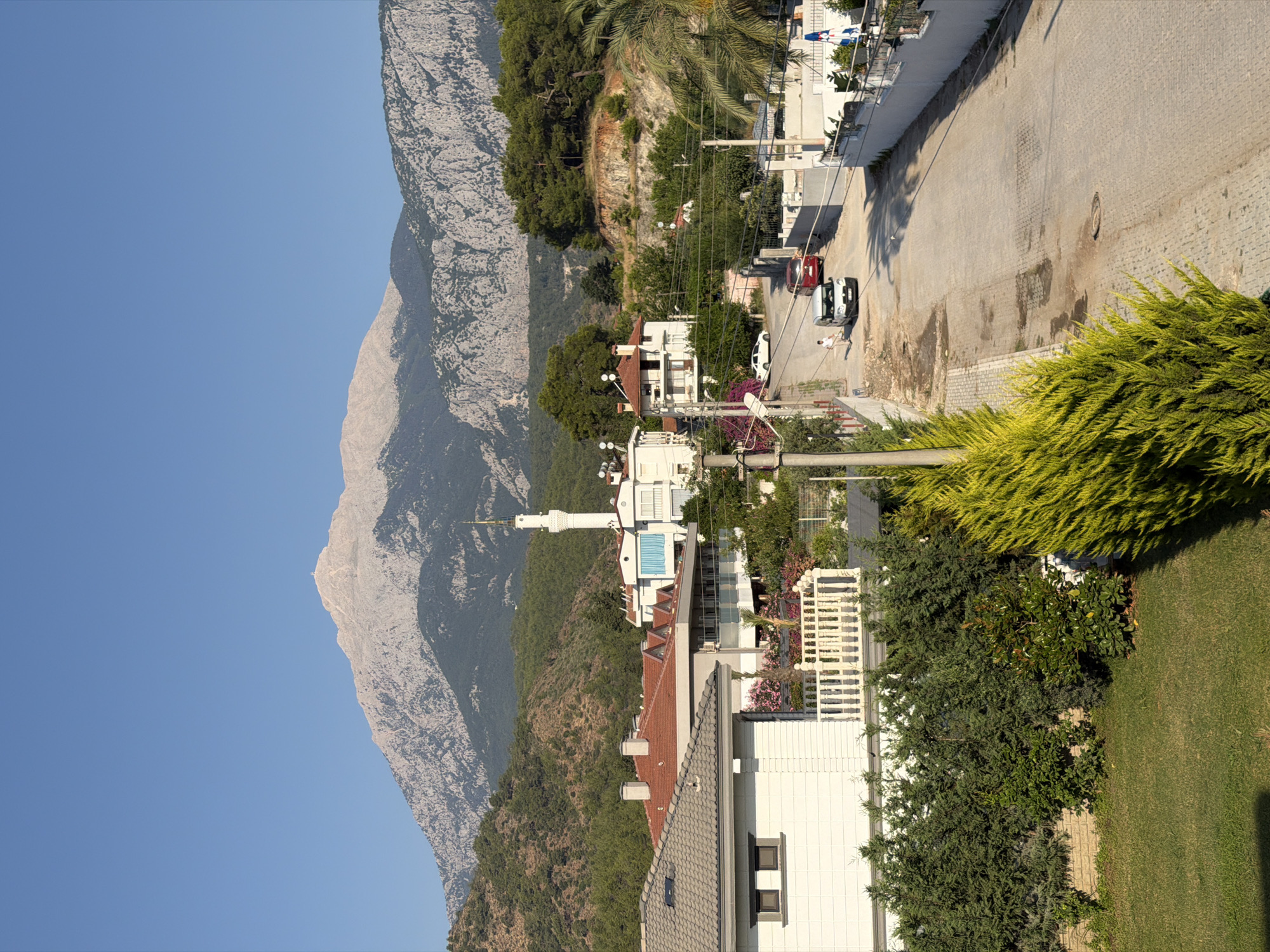 This is a vertical, eye-level, medium shot of a residential street in a hilly area with a large mountain range in the background. The foreground features a manicured lawn and lush green shrubbery, including a vibrant lime-green evergreen in the right foreground. A paved road runs through the middle ground, curving slightly to the right and uphill. Several white and red cars are visible on the road. To the left of the road, there are white houses with red roofs, and further up the street, a white building with a tall, slender minaret rises into the blue sky. The background is dominated by a large, rugged mountain range, its peaks illuminated by sunlight, with dense green forests covering its lower slopes. The sky is clear and blue.