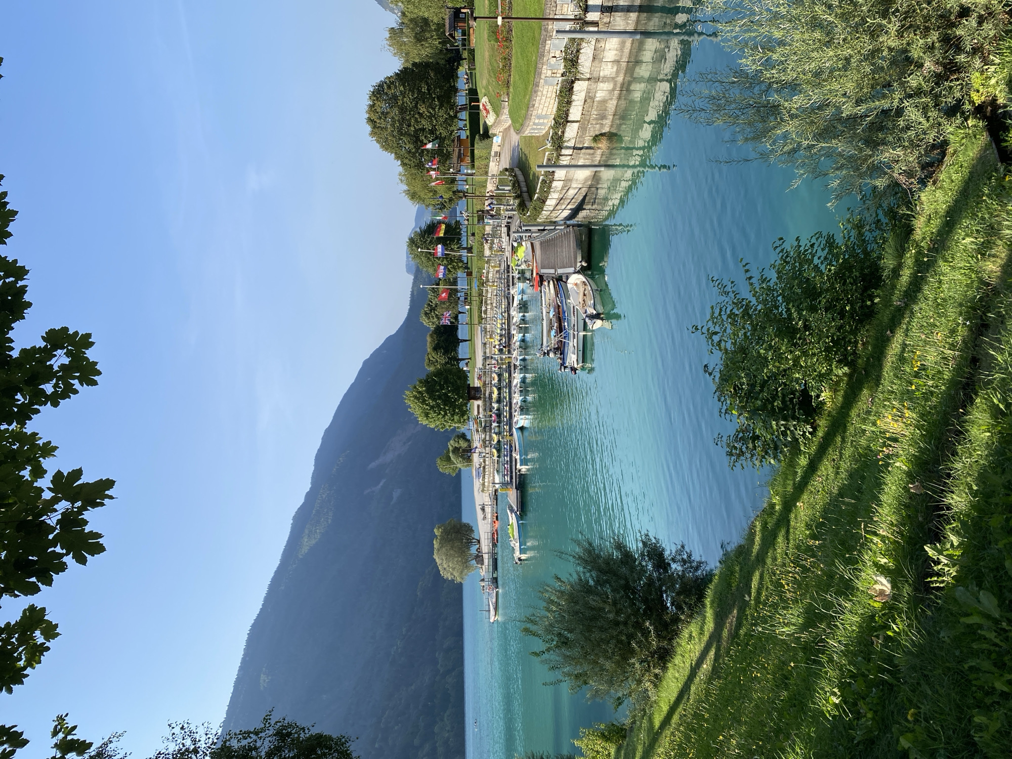 The image depicts a scenic view of a marina on a clear, sunny day in Molveno, Italy. In the foreground, a lush green grassy slope is illuminated by sunlight, with shadows cast by unseen trees. The slope leads down to the edge of a body of tranquil, turquoise water.
The water is clear and reflects the sky and surrounding landscape. A wooden pier extends into the water, lined with numerous small boats moored alongside. Some of these boats are dinghies, while others appear to be small motorboats. A few people can be seen walking on the pier and near the boats.
Beyond the marina, a sandy beach is visible, with a few scattered chairs and umbrellas. Further in the background, the landscape rises into gently sloping, tree-covered mountains under a clear blue sky. The trees in the foreground and along the shoreline are vibrant green. Several flags are displayed on poles along the waterfront, adding a touch of color to the scene. The overall atmosphere is peaceful and idyllic, characteristic of a popular lakeside tourist destination.