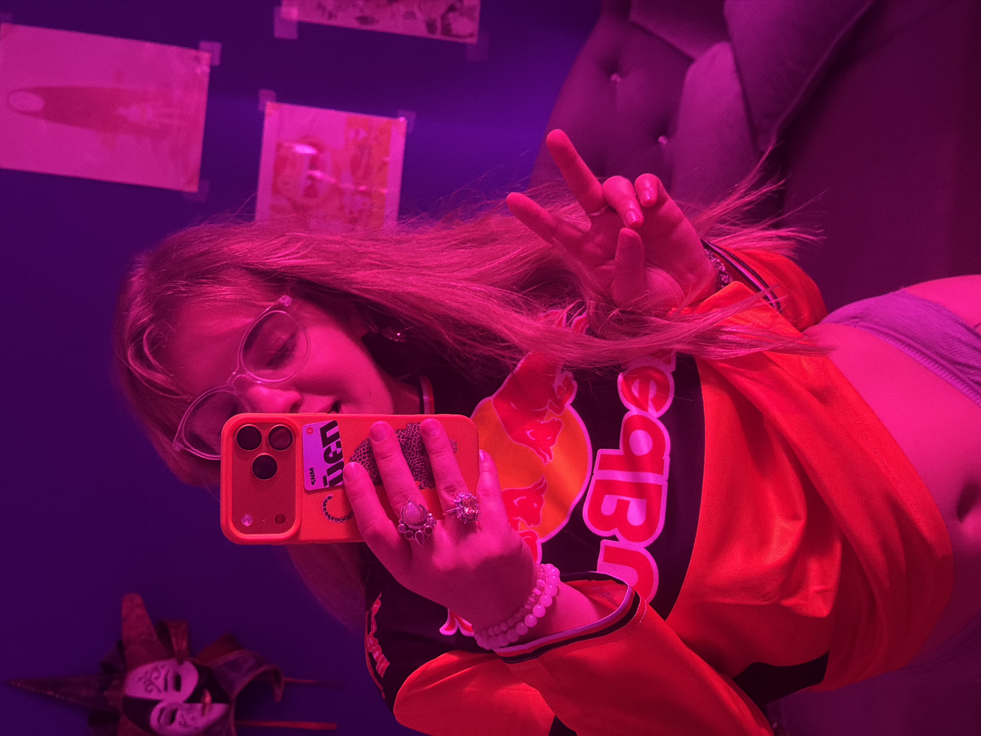 A person is taking a selfie in what appears to be a dimly lit room with a magenta/purple hue. The person is wearing glasses and an orange, black, and white jacket with the Red Bull logo prominently displayed. They are holding an orange iPhone with a sticker on the back. The person is making a peace sign with their fingers. There are rings on their fingers and a beaded bracelet on their wrist. A decorative mask is visible in the reflection below. On the wall are several papers or posters attached with tape. There is a tufted headboard visible in the background. I cannot confirm the specific location in Большая Шильна, Russia from this image.