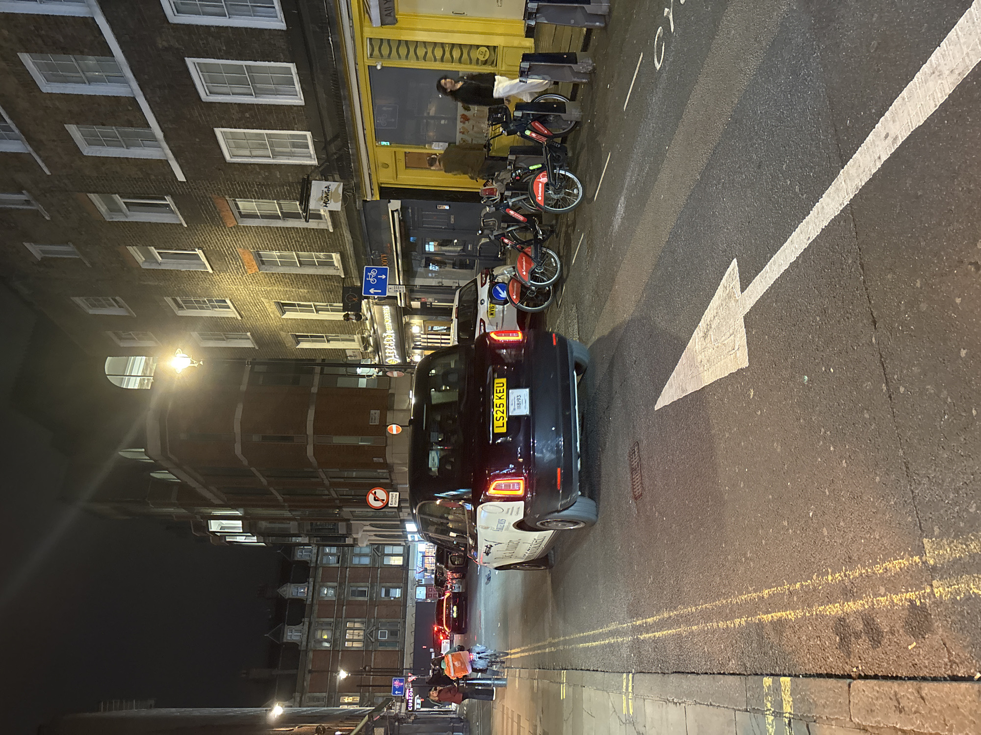 The image depicts a street scene at night in London, United Kingdom. A black taxi with license plate "LS25 KEU" is parked on the right side of the road, partially blocking a white arrow painted on the road surface. Several Santander Cycle rental bikes are parked along the left side of the road. A person stands near a shopfront with yellow framing. In the distance, pedestrians are visible on the sidewalk. The buildings in the background are illuminated by streetlights, and various street signs are also visible. The road surface appears wet, reflecting the ambient light.
