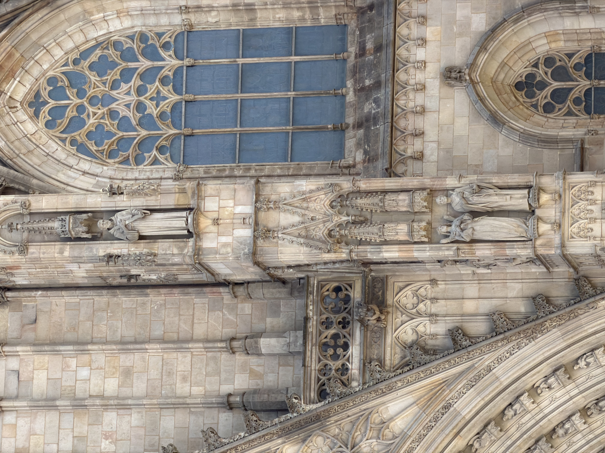 This is a close-up, overhead view of the ornate stonework of a Gothic cathedral, likely in Barcelona, Spain, given the location context. The image showcases intricate architectural details.
Prominently featured are two large stained-glass windows. The left window is a pointed arch shape filled with tracery, creating a delicate, floral-like pattern of stone divisions. Behind this tracery, the glass appears to be a deep blue. The right window is a smaller, similarly styled circular or quatrefoil window, also with blue glass.
Below the windows, there are several niches containing carved stone figures. Two figures are clearly visible, depicted in flowing robes, appearing to be saints or religious figures. They are positioned in shallow alcoves, and their details suggest a classical or medieval style.
The facade is constructed of light-colored, possibly sandstone, blocks arranged in a regular pattern. There is extensive use of carved ornamentation, including decorative moldings, cornices, and pinnacles, typical of Gothic architecture. A prominent feature is a large, curved architectural element, possibly part of an arch or vault, with intricate carved patterns and decorative friezes. The stonework shows signs of age and weathering, with some areas appearing darker or more worn than others.
The lighting suggests it is daytime, with the sun casting subtle shadows that highlight the three-dimensionality of the carvings. There are no people or obvious activity depicted, focusing solely on the static architectural elements of the building. The scene is one of grandeur and detailed craftsmanship, characteristic of a significant historical religious structure. No visible text is present in the image.