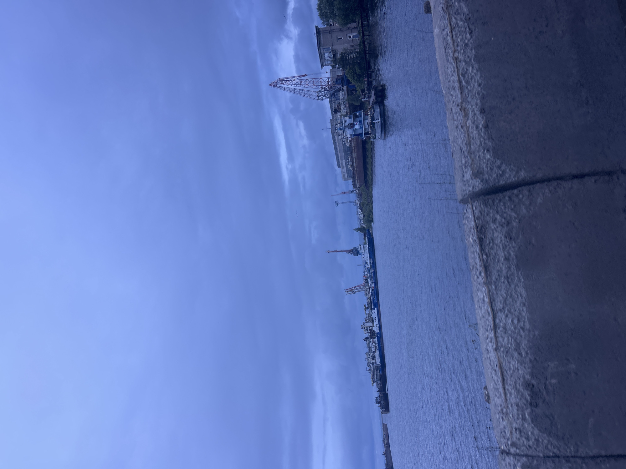 This image depicts an industrial waterfront scene in Chaikovsky, Russia, under an overcast, dark blue sky. A wide body of water, likely a river, dominates the midground, with a textured, dark concrete barrier visible in the blurred foreground, suggesting the photo was taken from a bridge or embankment.
On the far bank, several large industrial vessels, including what appear to be ships or barges, are moored or docked. Multiple large cranes, some red and some grey, are visible along the shoreline, indicating a shipyard or port facility. Industrial buildings line the bank behind these vessels, interspersed with some vegetation.
On the closer bank, to the right, a white and blue tugboat-like vessel is docked at a concrete pier. Immediately behind it, a prominent red lattice boom crane stands tall. An adjacent multi-story concrete building with windows, possibly an operations or administrative structure, is also visible. Further along this bank, more industrial structures and cranes are present, along with some trees and foliage. No individuals are discernible in the frame. The overall impression is an active but currently quiet industrial setting under subdued lighting.