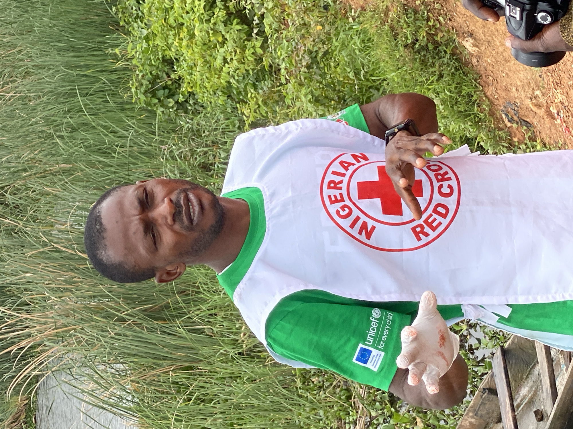 A Black male, wearing a green t-shirt with a UNICEF logo and a white vest with the Nigerian Red Cross emblem, stands in front of tall green grass and vegetation. He is speaking and gesturing with his hands, one of which is covered in a white glove with red markings. In the foreground, a person's hands are holding a camera. The scene appears to be outdoors, possibly near a body of water, with a wooden boat to the left of the frame. The overall impression is of a humanitarian aid worker engaged in their duties in a natural setting.