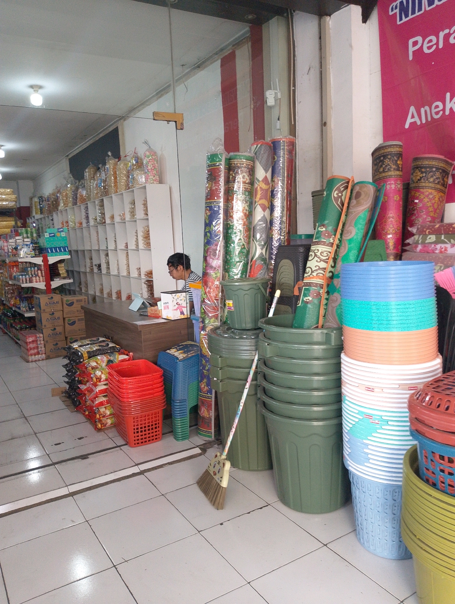 This image depicts the interior of a store located in Lembang, Indonesia, likely selling a variety of household goods and snacks. In the background, shelves are filled with packaged snacks. A person wearing glasses and a striped shirt is seated at a wooden counter, looking at a tablet. In the foreground, there are stacks of red plastic baskets, blue plastic stools, and various sizes of green plastic buckets. To the right, there are stacks of large plastic containers in blue, light blue, and orange. A broom leans against a green bucket. Several rolled-up mats with patterned designs are also visible. The floor is tiled with off-white tiles.