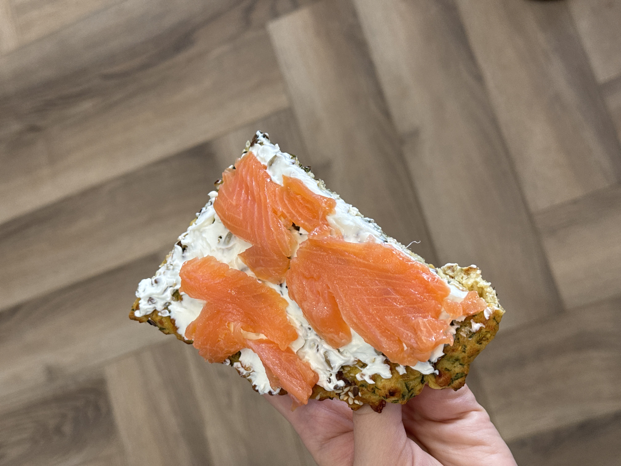 Here's a description of the image:
The image shows a close-up of a piece of food held in someone's hand. The food appears to be a savory pancake or bread topped with cream cheese and smoked salmon. The base has a slightly browned, irregular edge and seems to contain seeds or grains. The cream cheese is spread evenly over the base, and several slices of smoked salmon are artfully arranged on top. The person holding the food is only partially visible, with a glimpse of fingers. The background is a wooden floor with a herringbone pattern. The photo is taken indoors, with natural lighting.