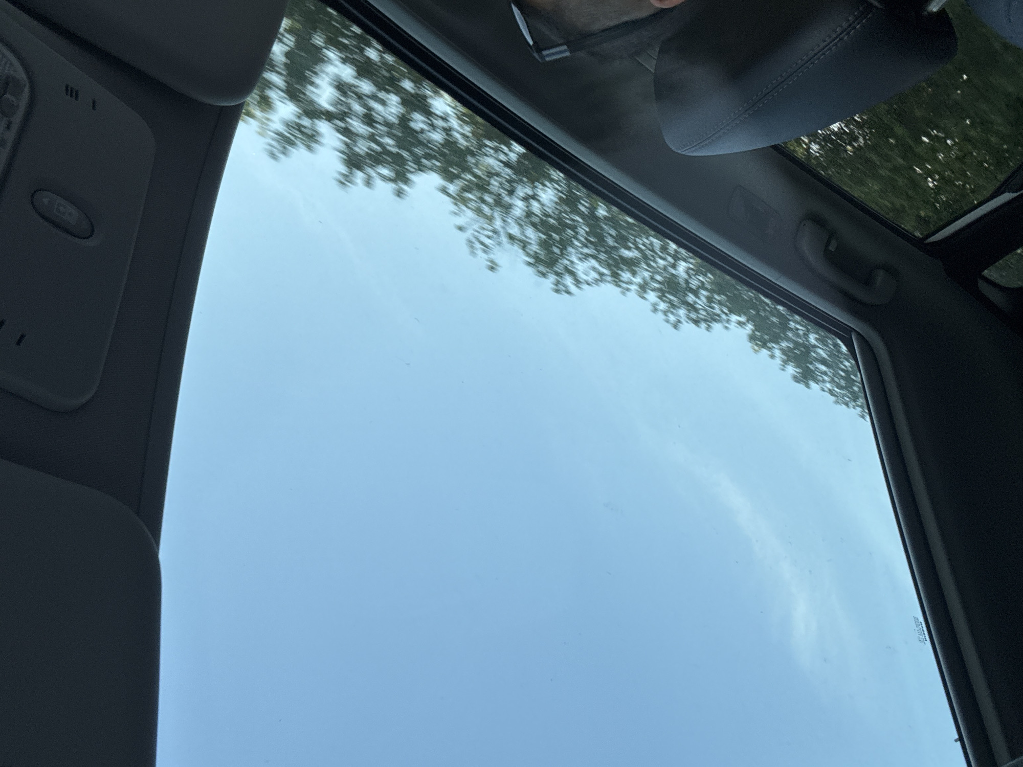 The image shows a view from inside a car, looking up at the sky through a sunroof. The sky is a light blue, with some faint wispy clouds. Trees are visible outside the car, blurred by motion, suggesting the car is moving. The interior of the car is dimly lit, with parts of the headliner and a sunroof control panel visible at the top. A headrest of a car seat is visible on the right side of the frame, and a person's head and shoulder wearing glasses can be seen in the upper right corner. The scene appears to be a drive through a tree-lined area, possibly in or near the city of Cojușna, Moldova.