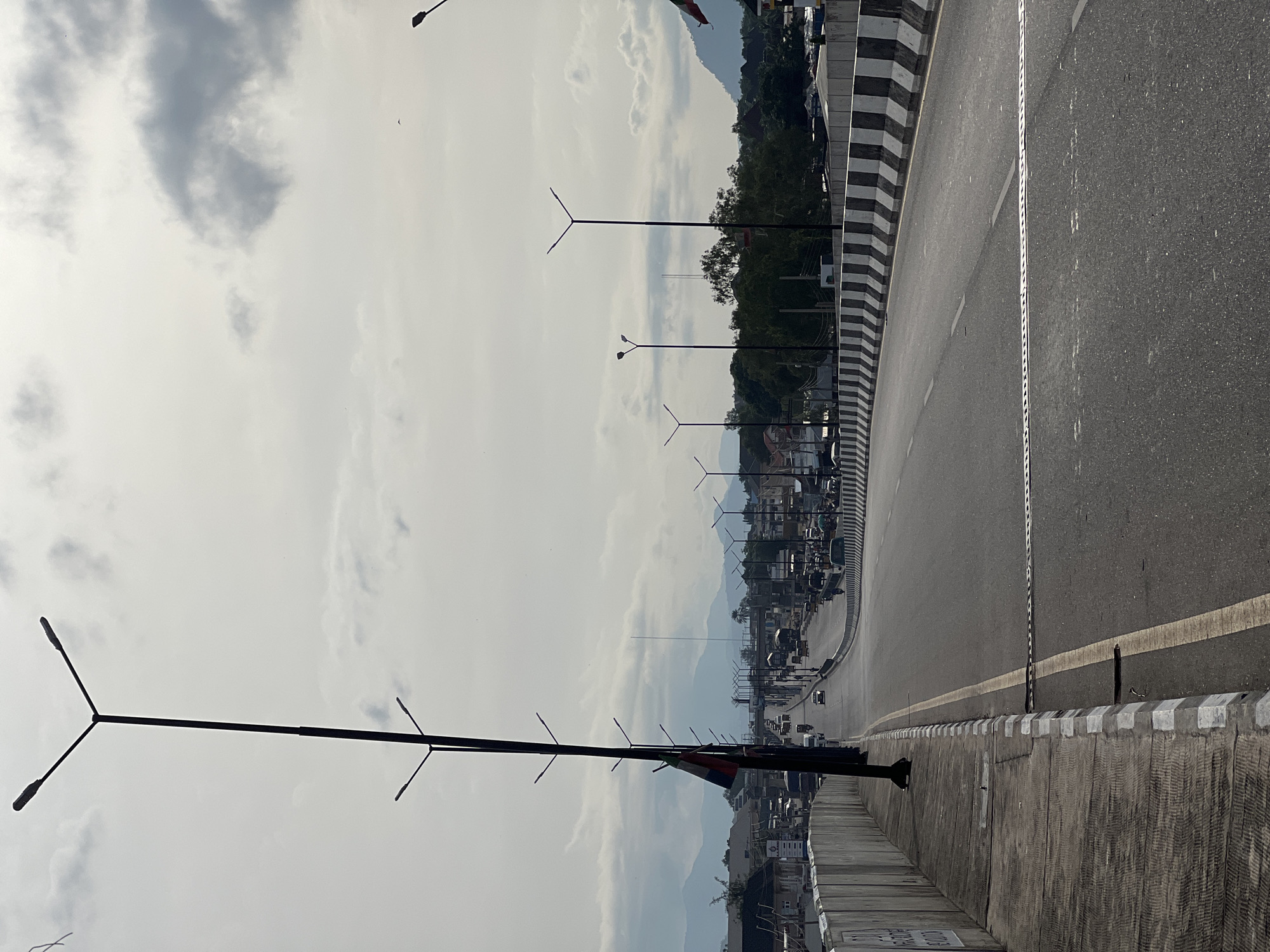 The image shows an overpass or bridge in Jalingo, Nigeria, with traffic and a cityscape visible in the background. The perspective is from the side of the road, looking down the length of the bridge.
On the left side of the image, there is a concrete walkway with a textured surface and a white and grey striped border. A tall black lamppost with two lights extends upwards from this walkway. A flag, appearing to be green, white, and red, is attached to this lamppost. Further along the walkway, more lampposts are visible, spaced out along the edge of the bridge.
The road itself is asphalt and is divided into lanes by white lines. There are several cars, trucks, and motorcycles on the road, indicating moderate traffic. The road curves gently to the right as it recedes into the distance.
On the right side of the bridge, there is a concrete barrier with alternating black and white stripes. Beyond this barrier, the cityscape of Jalingo can be seen, with buildings of various sizes and shapes. Green trees are interspersed within the urban environment. In the far distance, mountains are visible under a cloudy sky. The sky itself is overcast with large grey clouds.
The overall scene suggests an active urban environment on a bridge, with typical road infrastructure and elements of the surrounding city and natural landscape.