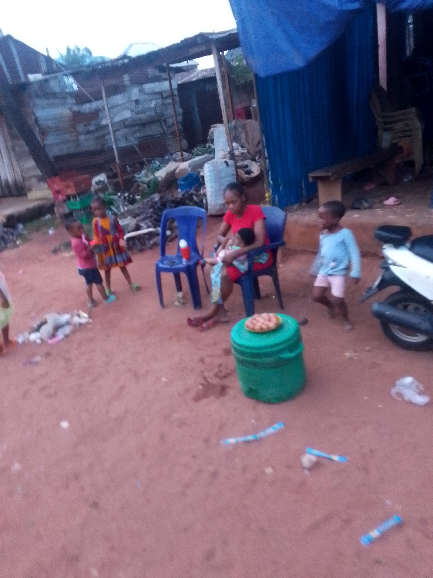 The image depicts an outdoor scene in what appears to be a residential or semi-commercial area in Orlu, Nigeria. The time of day is likely late afternoon or early evening, characterized by soft, diffused light, suggesting an overcast sky or twilight.
The central subjects are an adult woman and several children gathered on a reddish-brown dirt ground. The woman, wearing a red top, is seated on a blue plastic chair, holding a baby or toddler on her lap. Several other children are standing around; one child in a light blue long-sleeved shirt and pink shorts walks towards the right, while another in a pink top and blue shorts and a third in a colorful patterned dress are visible to the left.
In the immediate foreground, a green cylindrical cooler or container is present, with a plate of small, round, baked goods or fritters placed on its lid. The dirt ground is littered with several blue and white wrappers, likely from snacks or candies. A white motorcycle is parked on the right side of the frame.
The background consists of makeshift structures, primarily constructed from corrugated metal sheets and wooden supports. A large blue tarp or awning covers part of a structure on the right, under which a wooden bench and stacked plastic chairs are visible. To the left, stacked red and green plastic crates can be seen. The area behind the figures appears somewhat cluttered with various items, including what looks like discarded bottles and other materials. There is no discernible text visible in the image. The overall setting portrays a casual, everyday outdoor gathering in a less formal environment.