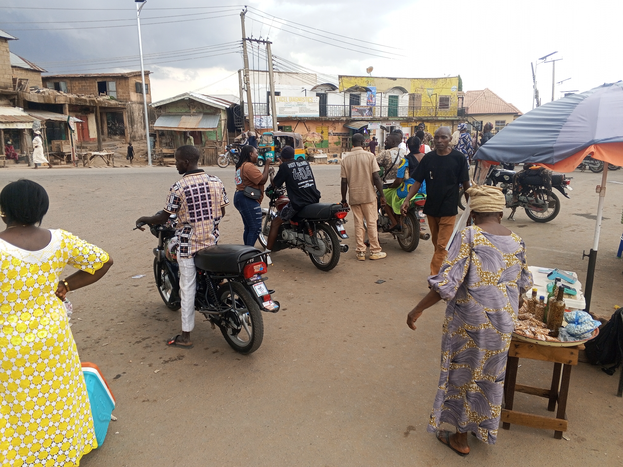 The image captures a street scene in Gwong, Nigeria. Several people are present, some standing and interacting, others on motorcycles. A woman in a yellow and white dress stands with her hand on her hip, while another woman in a purple and gold patterned dress is near a small table displaying items for sale. Several motorcycles are parked in the center of the frame. Buildings are visible in the background, some appearing to be shops or commercial spaces. The sky is overcast, casting a diffused light on the scene.
