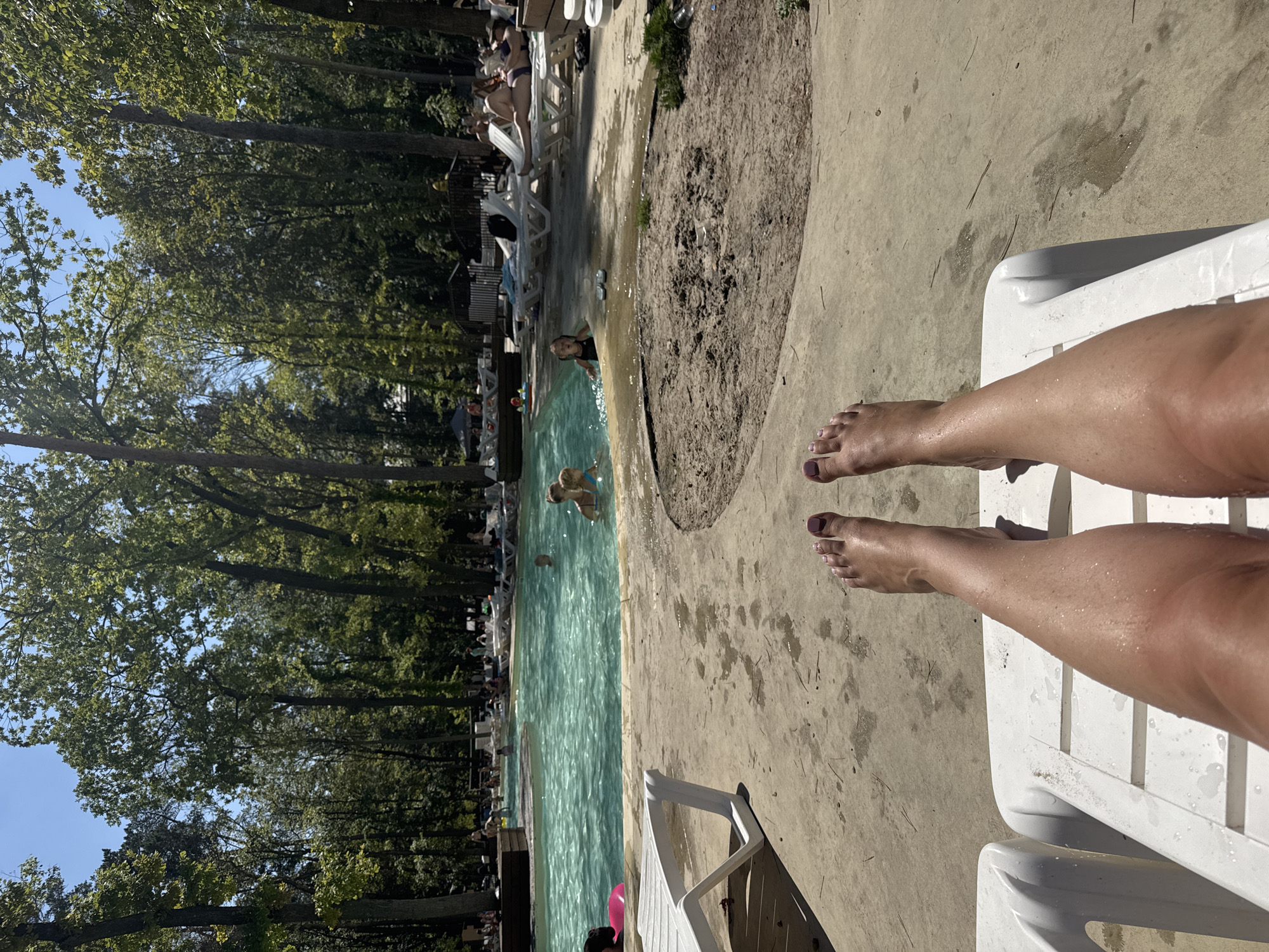 The image depicts a sunny day at an outdoor swimming pool complex located in the city of Orlivshchyna, Ukraine. The perspective is from a person lying on a white lounge chair, with their legs and feet in the foreground, indicating they are relaxing poolside. In the middle ground, a bright blue pool is visible, with several people, including children, swimming and playing in the water. The pool area is surrounded by concrete walkways and more white lounge chairs. Beyond the pool, a dense area of tall green trees creates a natural backdrop. Several people can be seen relaxing on lounge chairs under umbrellas or enjoying the pool. The environment appears to be a resort or recreational facility.