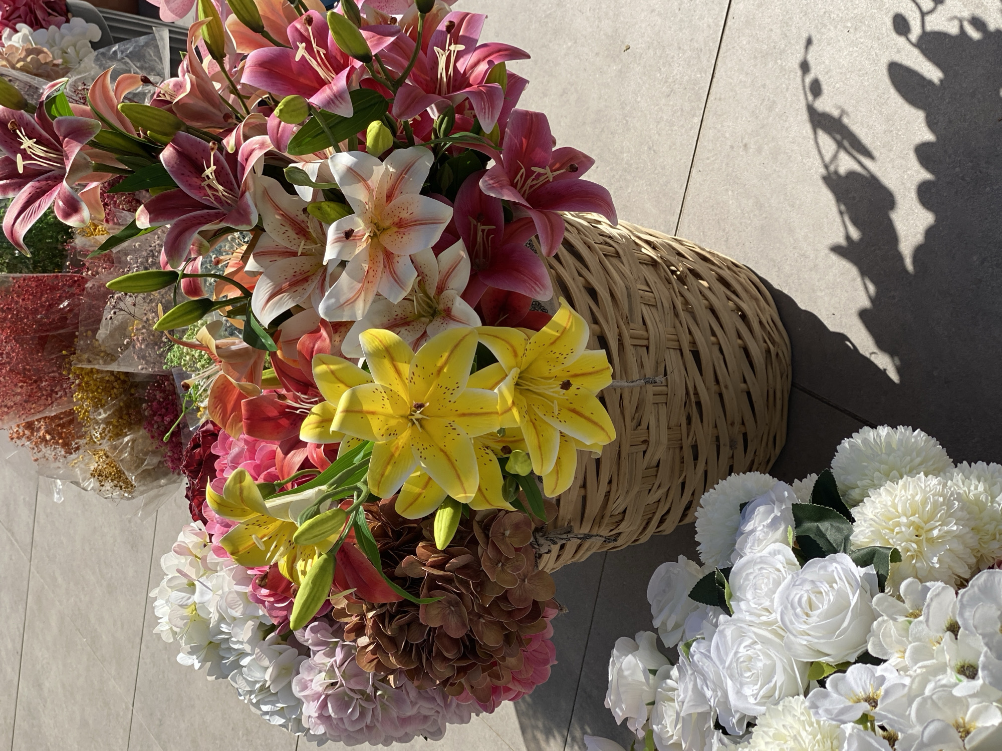 The image shows an assortment of artificial flowers in a woven basket, and other bouquets. The dominant flowers are lilies in pink, white with pink speckles, and yellow. There are also white roses and other flowers, plus brown hydrangea-like flowers. Some flowers are also arranged in cellophane. The scene appears to be outdoors, possibly a market stall. The location is Çekmeköy, Turkey. There is a shadow cast on the tile surface on the right side of the image.