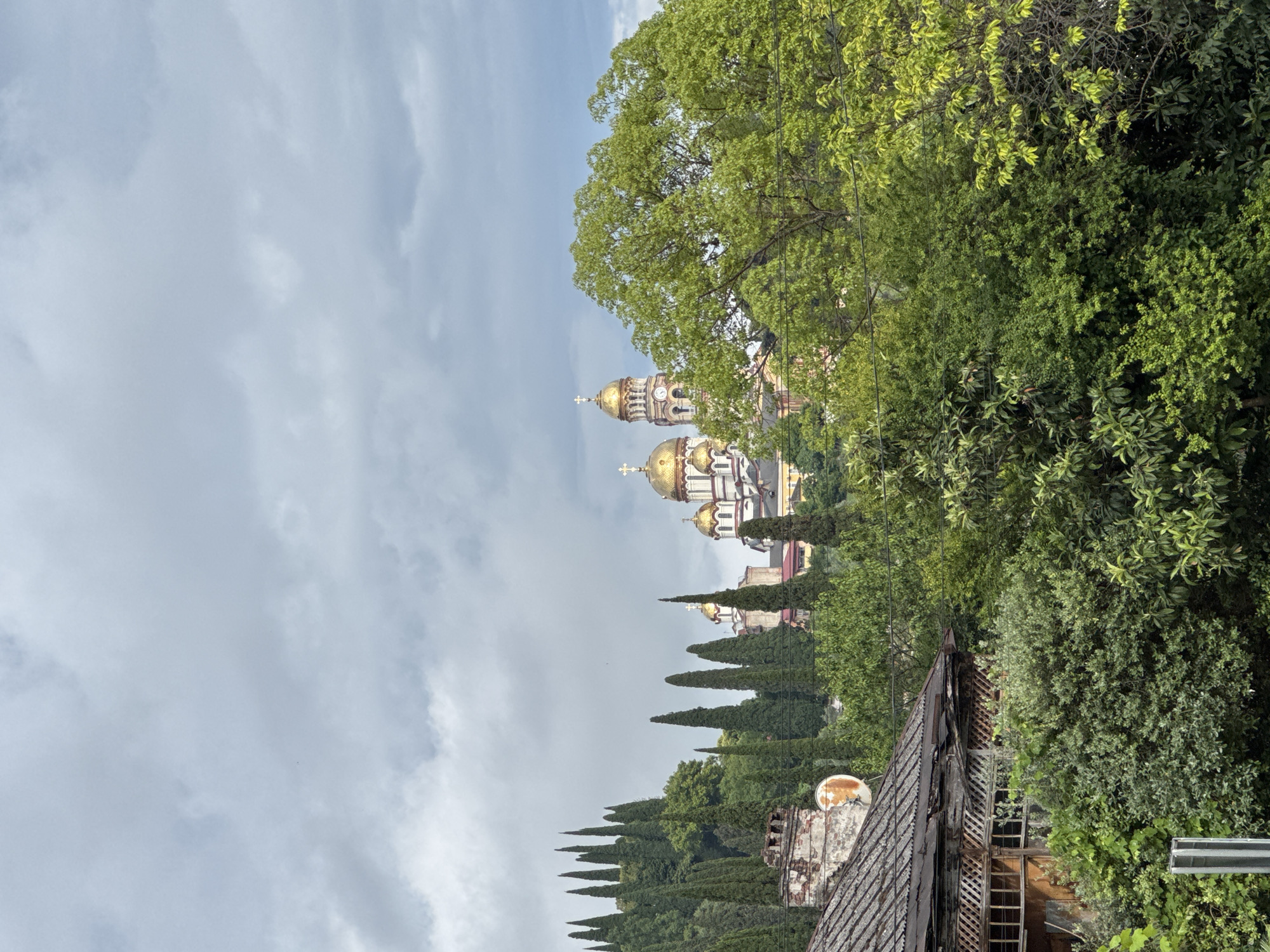 The image depicts a scenic view of New Athos, Abkhazia, featuring the iconic New Athos Monastery with its golden domes prominently displayed against a backdrop of lush green trees and a cloudy sky. In the foreground, the weathered roof of an old building, possibly a house or outbuilding, with a rusted satellite dish on its chimney, adds a touch of rustic charm. Tall cypress trees line the hillside, leading the eye towards the grand monastery situated on the elevation. The overall atmosphere is serene and picturesque, capturing the natural beauty and architectural heritage of the location.