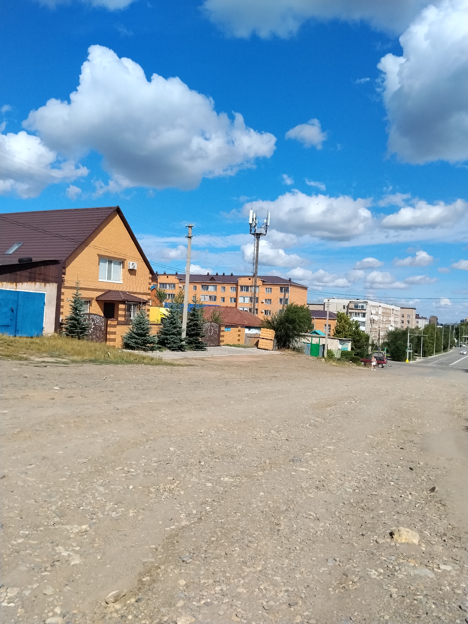 The image depicts an outdoor scene with a dirt road in the foreground. In the mid-ground, a two-story orange brick house with a brown roof is visible. Adjacent to the house are several small evergreen trees and a brown metal fence. Further back, there are multiple apartment buildings of varying heights and colors, including orange and white structures. A cellular tower stands tall against a bright blue sky with scattered white clouds. To the right of the apartment buildings, a paved road with vehicles and some greenery lines the side. A person is visible walking near a red car on the paved road. The overall environment suggests a residential area within a city. Based on the architectural style and the presence of trees, this scene is likely located in Kokshetau, Kazakhstan.
