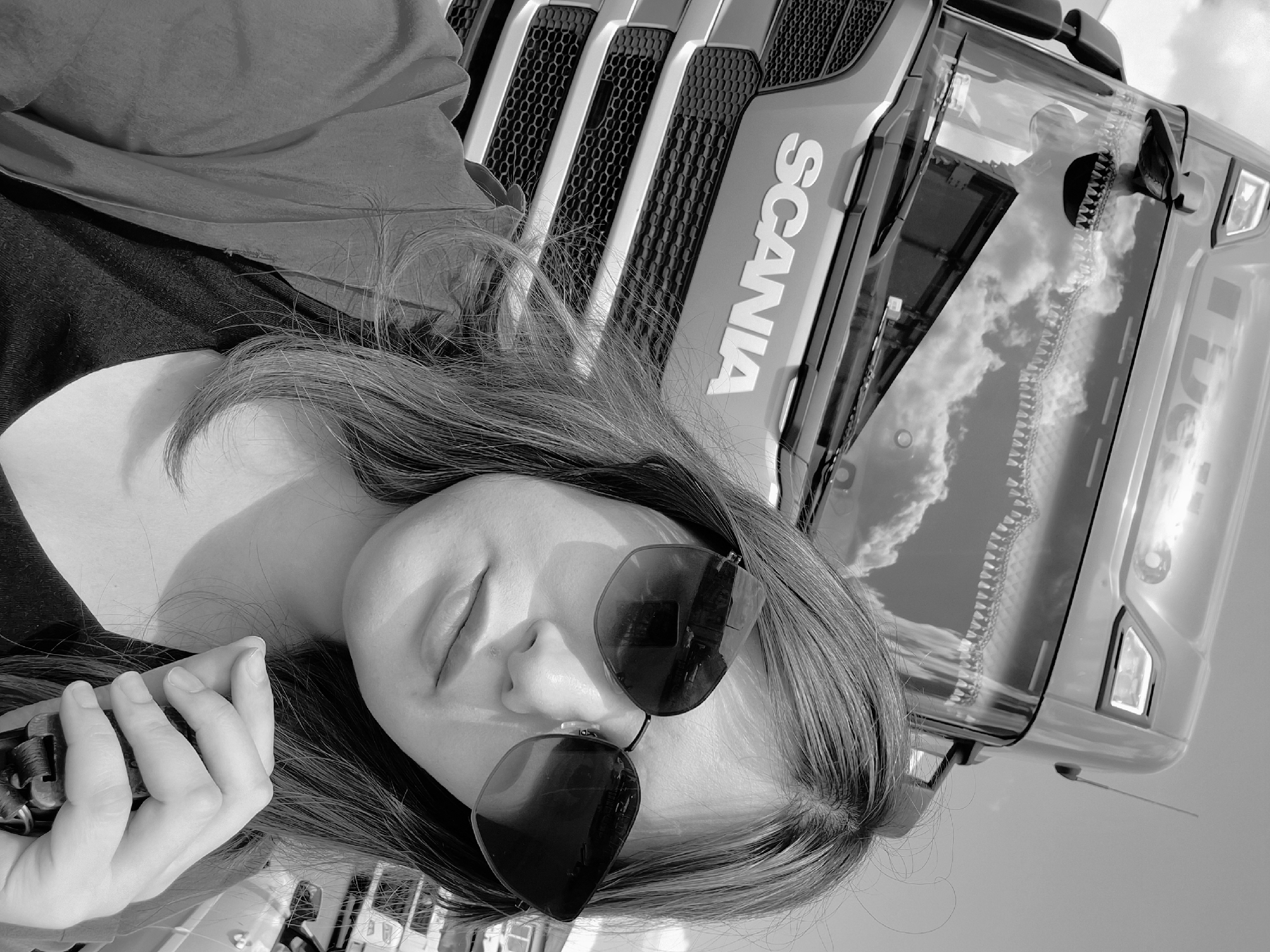 A black and white image displays a young person lying or leaning against the front of a large truck. The individual, seen from a low angle, has long hair and is wearing dark sunglasses, with their eyes appearing closed or cast downwards. They hold an unidentified object, possibly a mechanical component or a large clip, in their left hand.
The truck, a Scania, is prominent in the upper right portion of the frame. The "SCANIA" logo is clearly visible in white letters on the truck's grille. The windshield reflects a bright, cloudy sky, indicating a clear, sunny daytime setting. Reflections of distant structures or landscapes are also discernible in the truck's glass.
The scene takes place outdoors during the day. The person's demeanor seems relaxed or pensive. The context provided specifies the location as Izhevsk, Russia. The image is rotated, giving it a unique, dynamic composition.